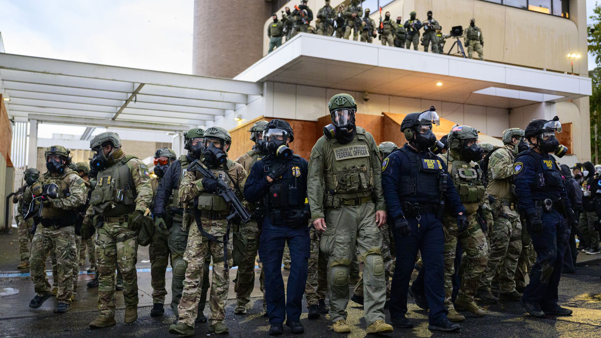 Group of armed officers and federal agents in tactical gear and gas masks standing in formation outside a building during dusk.