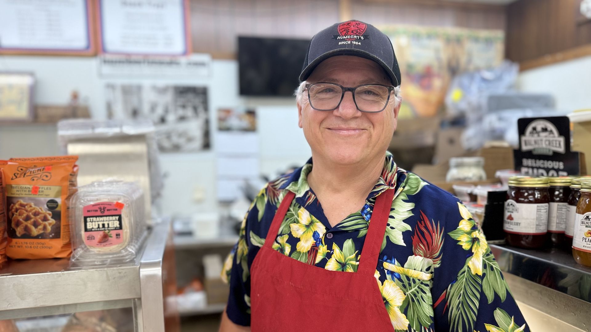 Smiling man in a red apron and black cap behind a deli counter, wearing a bright tropical shirt; shelves, jars, and snack packages visible in the background.