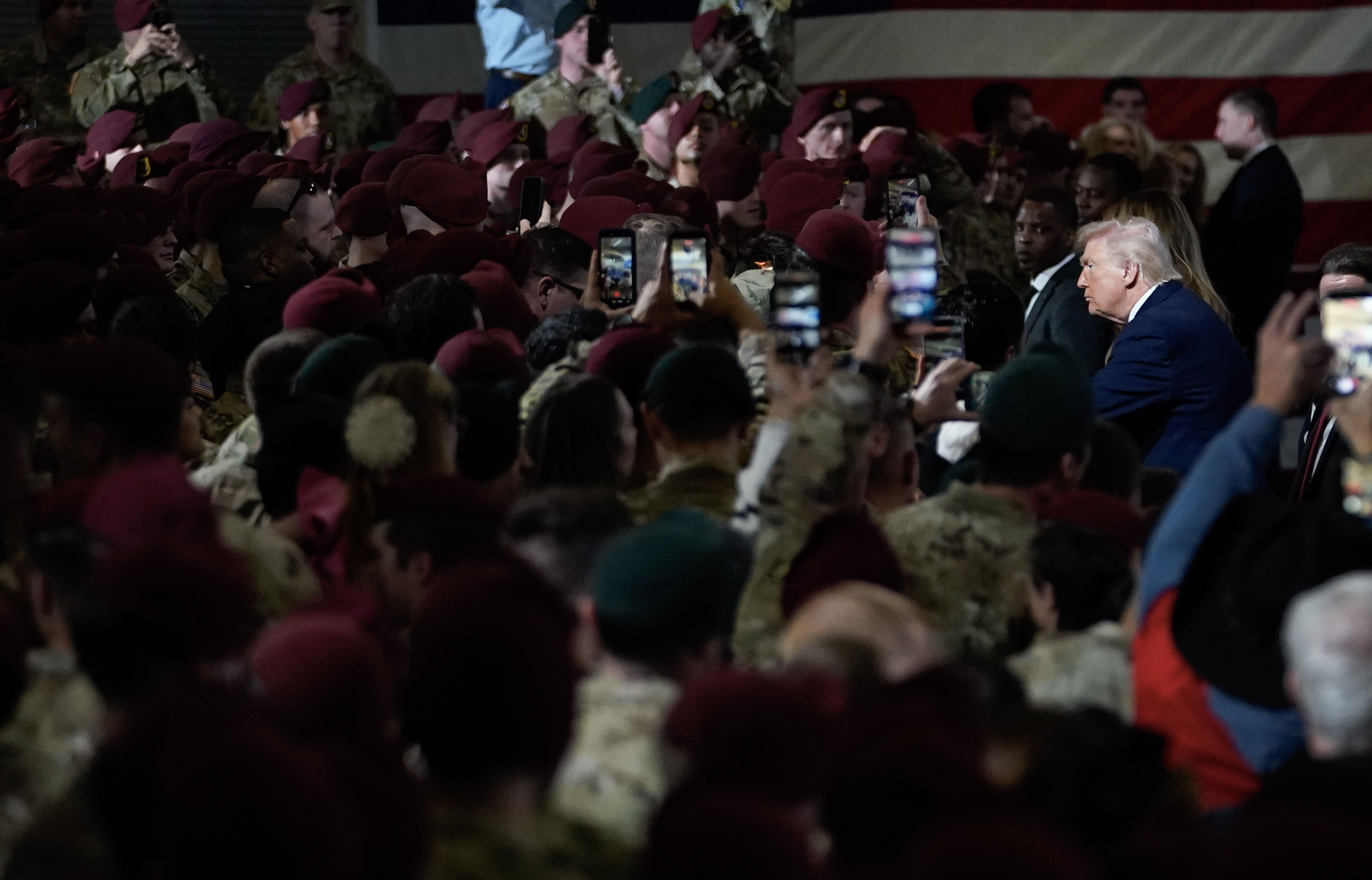 President Donald Trump in a blue suit speaks to a dense crowd of soldiers and civilians; many hold up phones to film, with a large American flag in the background.