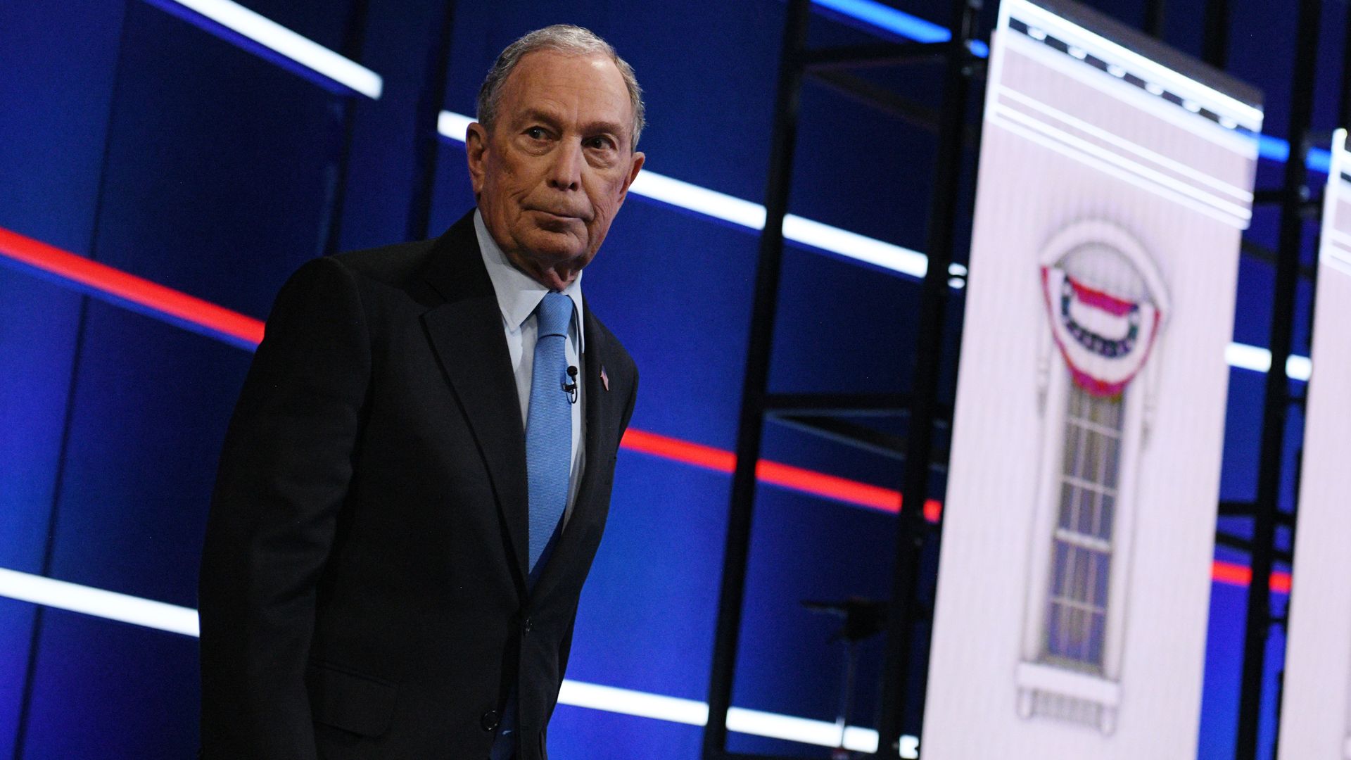 In this image, Bloomberg stands on the debate stage in a suit 