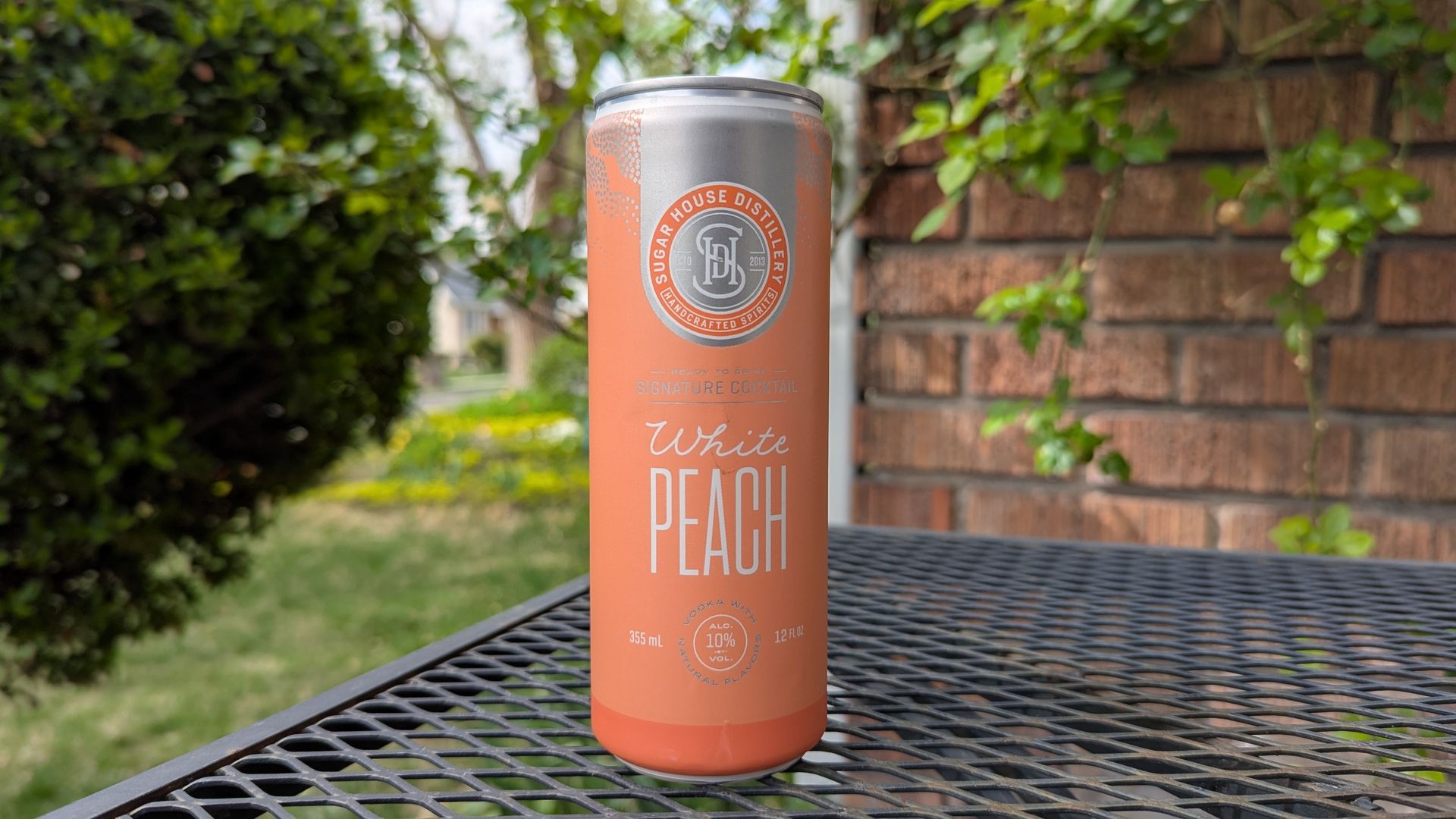 A peach-colored can says Sugar House Distillery White Peach. It sits on a black metal patio table, in front of a brick wall with vines and a green bush.