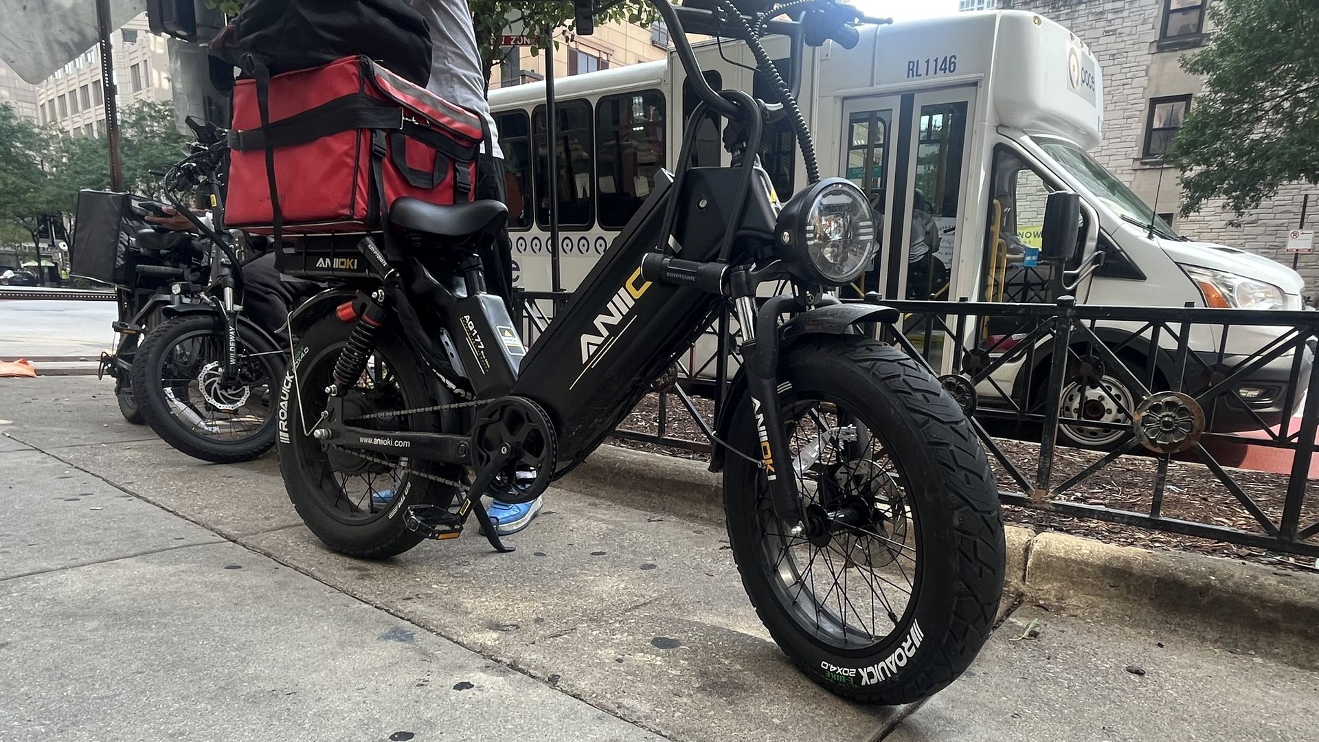 Black ANI electric bike with wide tires and a red insulated delivery bag attached to the back, parked on a city sidewalk near a white shuttle bus and buildings.