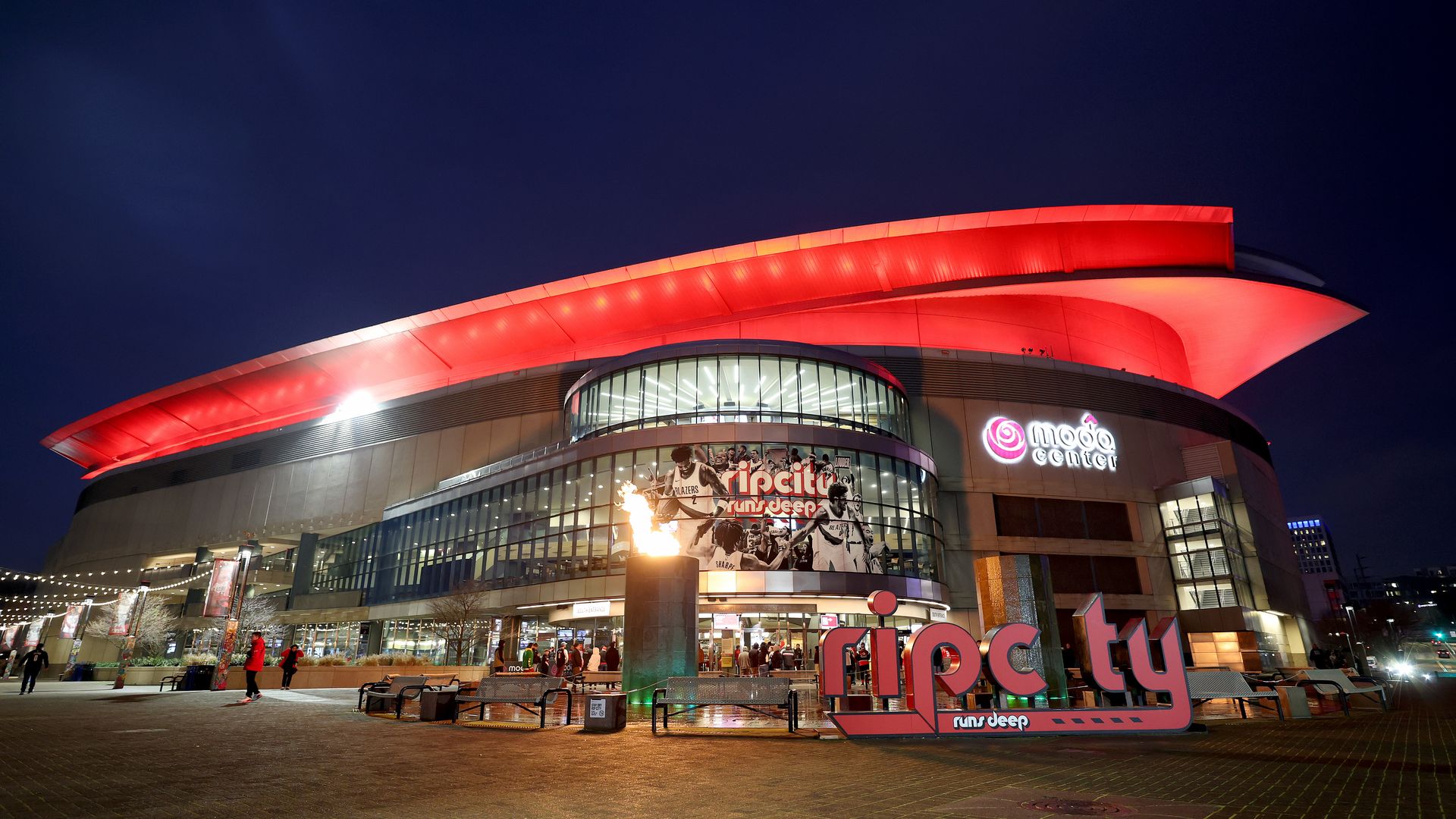 Nighttime view of a modern arena with a bright red curved roof, glass facade, and illuminated signs. People stroll the plaza under string lights as a dark blue sky looms.