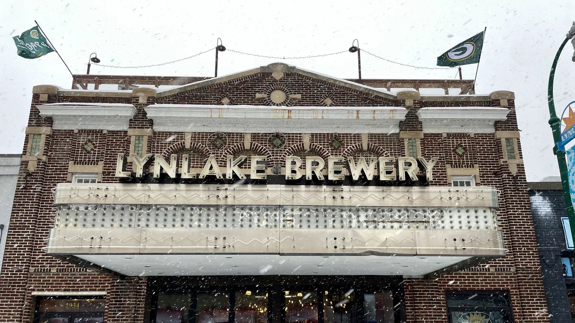 Brick building with the sign "LYNLAKE BREWERY" on a snowy day, Green Bay Packers flags flying on the roof, and a marquee below the sign.