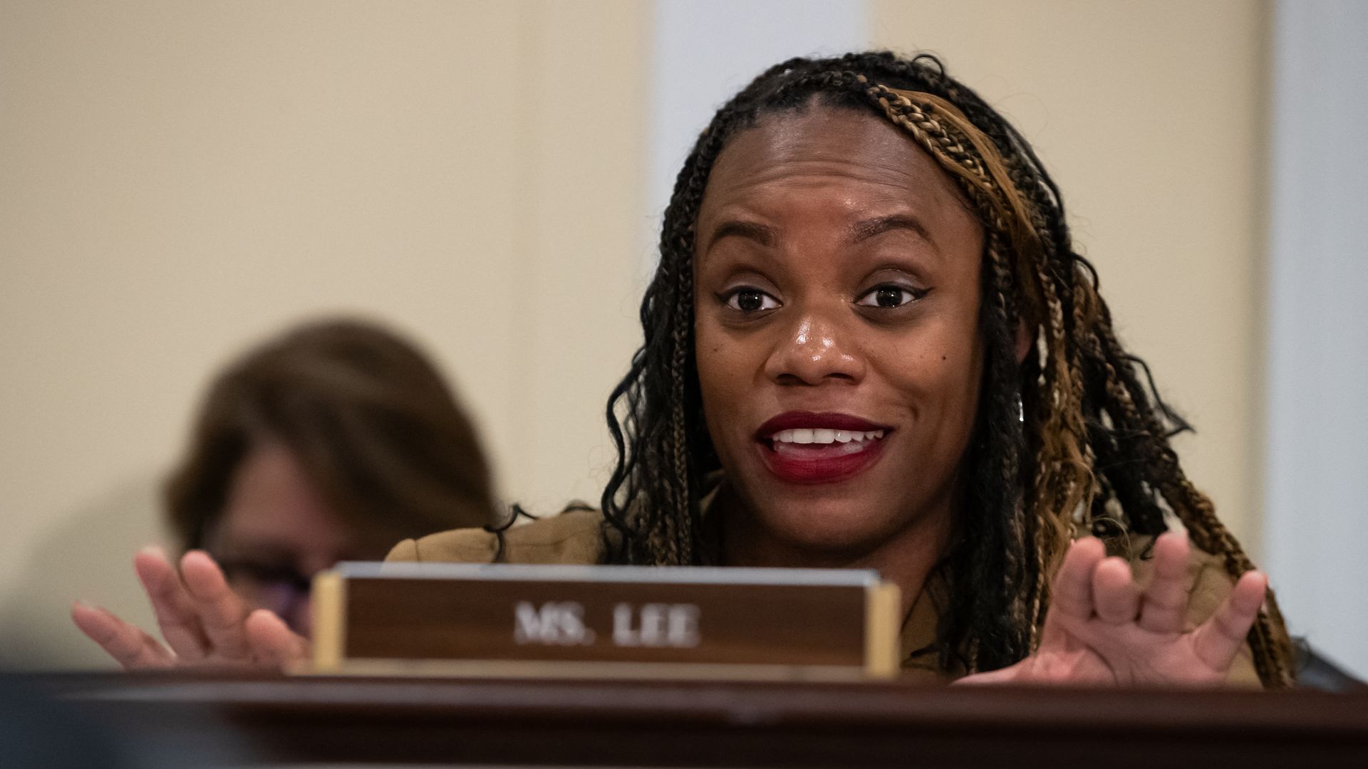 Woman with braided hair and red lipstick speaking behind a desk nameplate that reads "Ms. Lee," gesturing with her hands in a beige room.