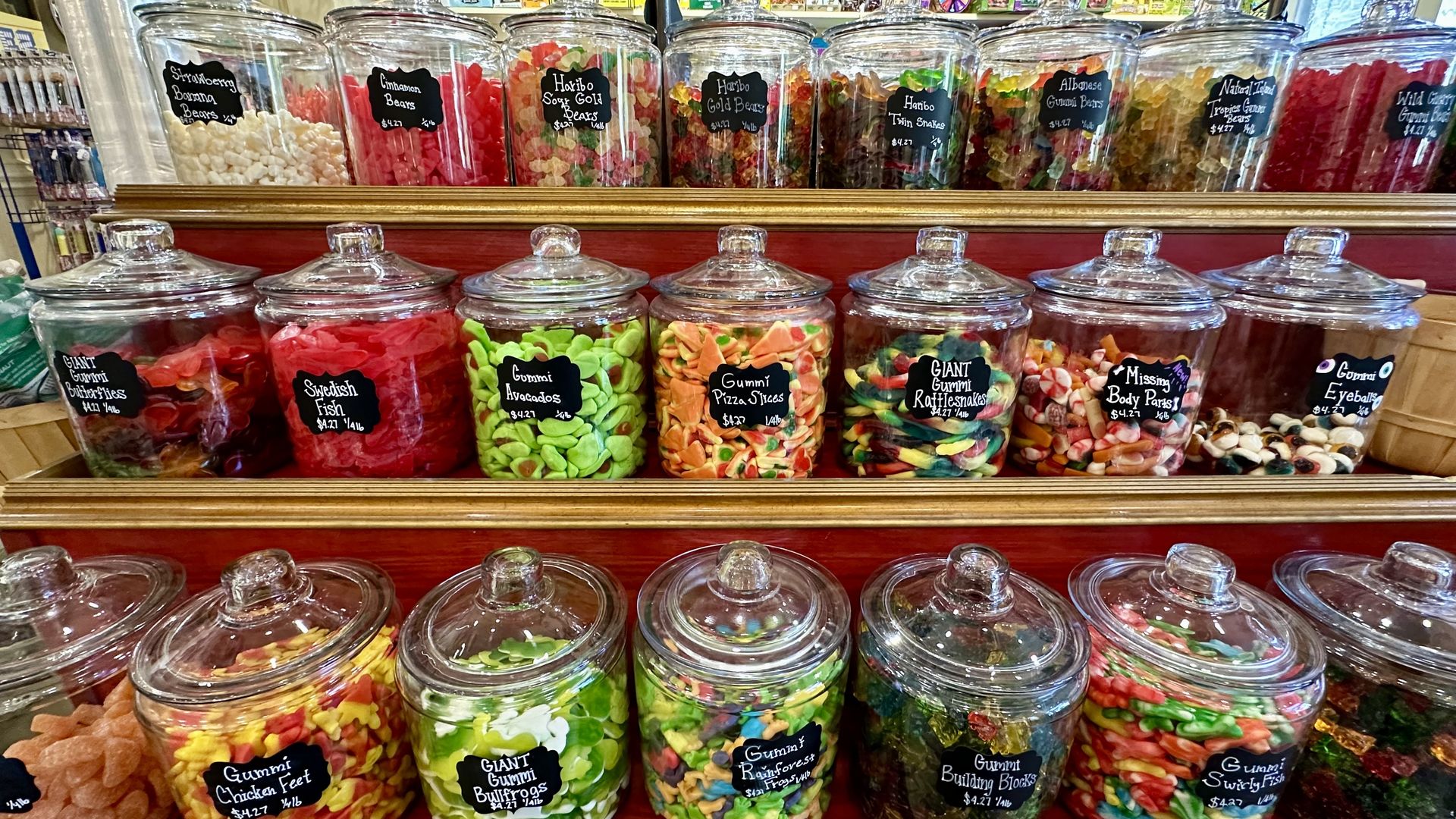 Row of clear glass candy jars with lids on wooden shelves, filled with colorful gummies (bears, worms, slices, fish). Black chalkboard labels read items like "Giant Gummi Bears" and "Swedish Fish".