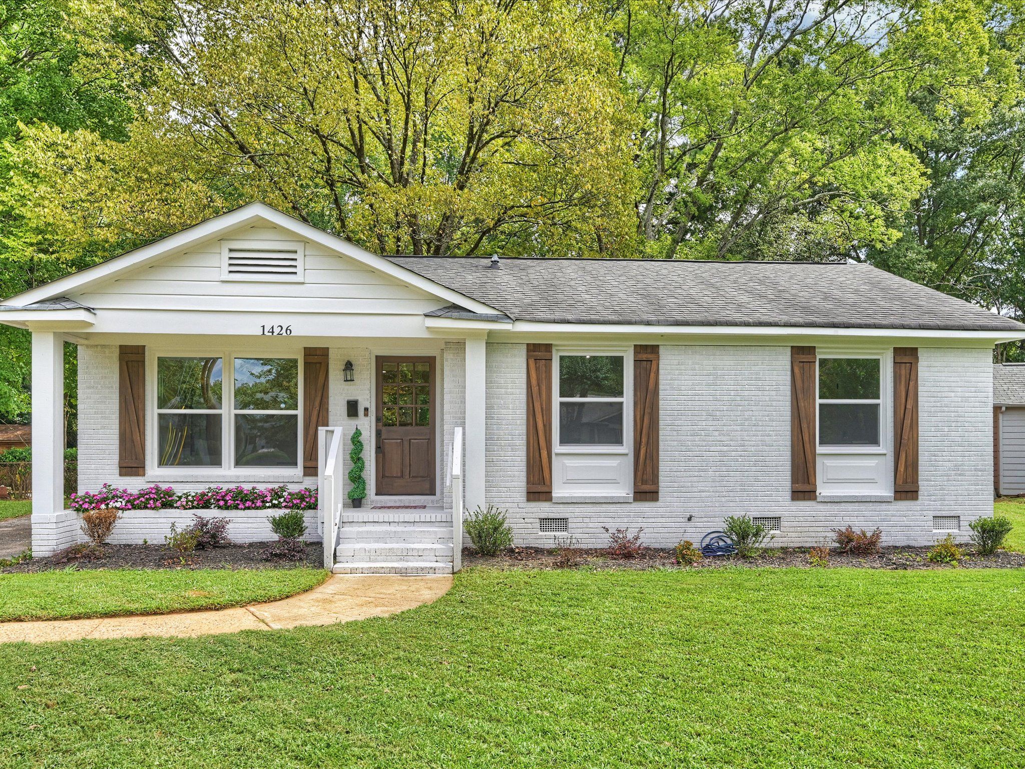 Single-story white brick house with brown wooden shutters, a brown front door, flower bed with pink flowers, and green lawn surrounded by tall leafy trees.