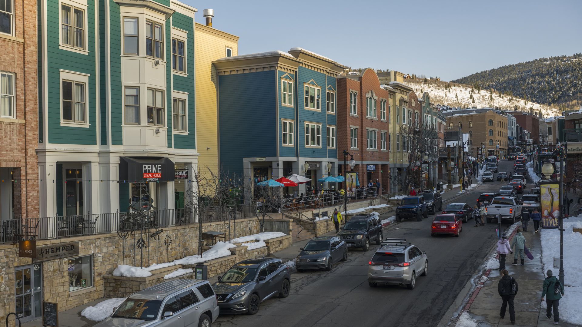 Winter street scene with parked cars and people walking near colorful buildings including a teal one with a "PRIME Steak House" sign, and snow-covered hills in the background under a clear sky.