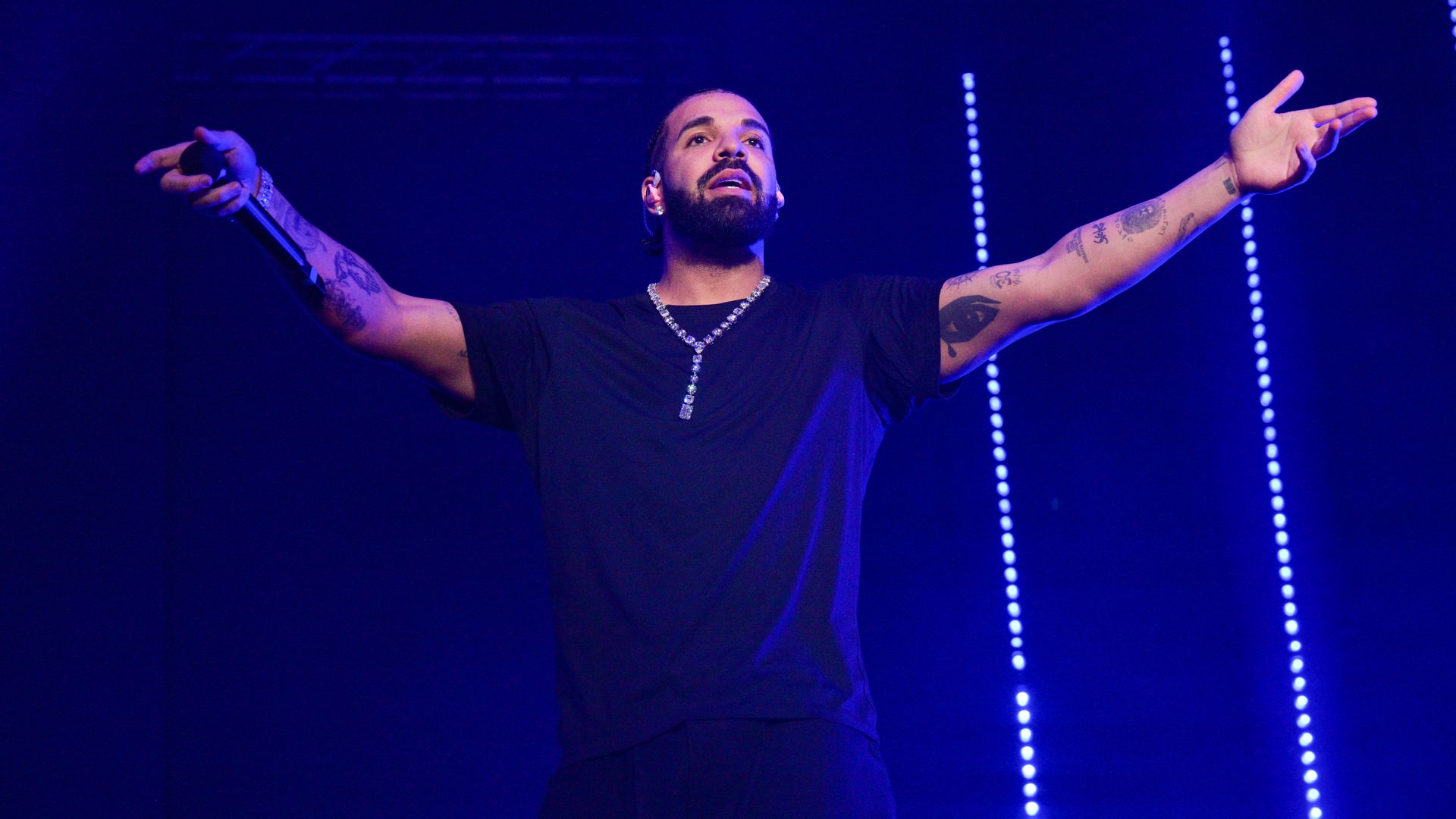 Drake seen on stage, in purple light, with his arms outstretched to the crowd.