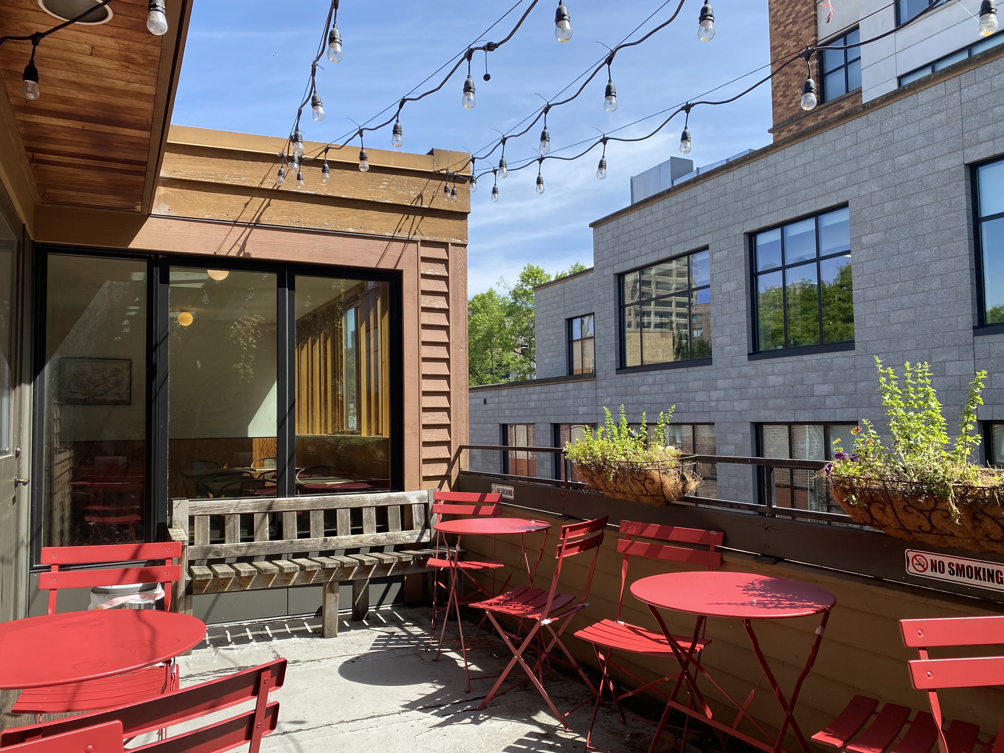 Red cafe tables on a deck with a building across an alley and a glass sliding door to reenter the cafe.