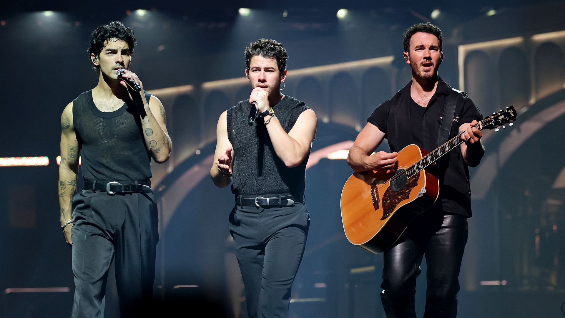 Three male singers perform on stage under bright lights; the man on the right plays an acoustic guitar, all wearing dark sleeveless or short-sleeve tops and black pants.