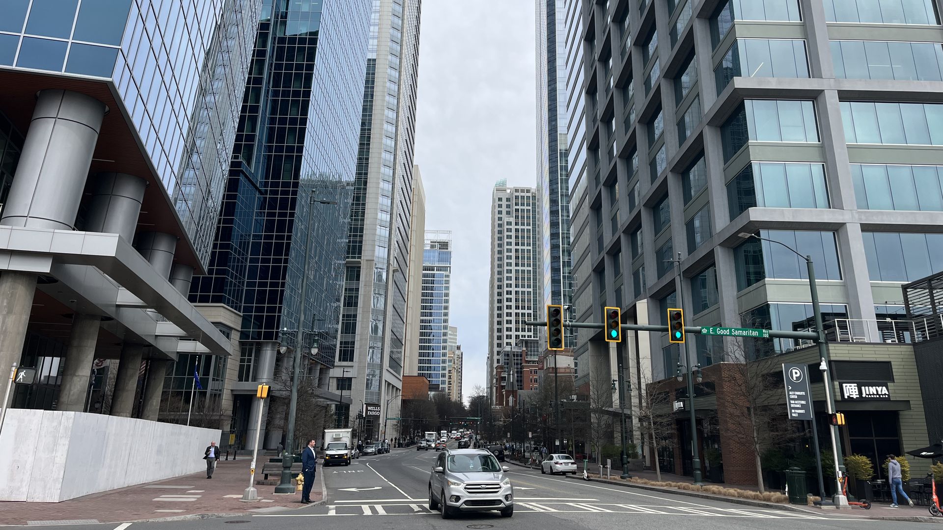 Urban street with towering glass and concrete skyscrapers on both sides. Cars on the road, traffic lights overhead. Pedestrians on sidewalks; a JINYA sign is visible on the right, under a cloudy sky.