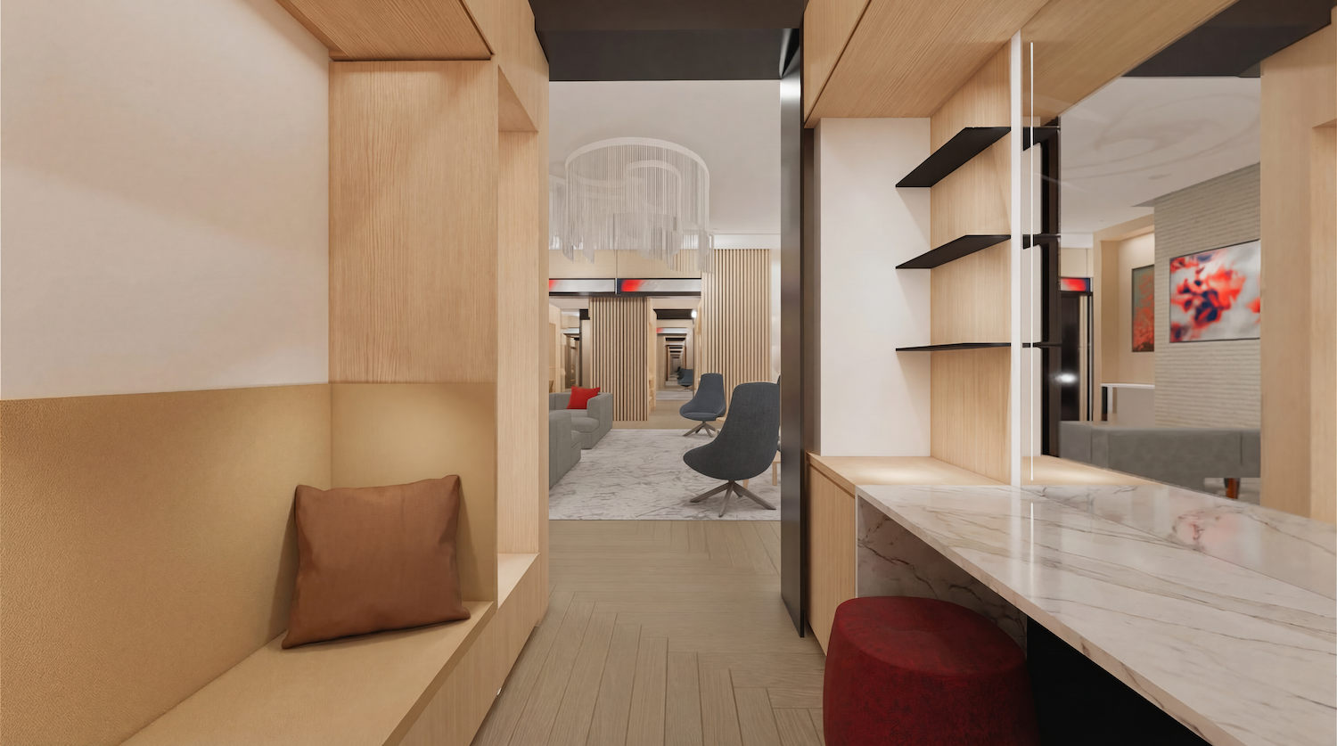 Modern hotel lounge seen through a hallway: warm wood panels, marble counter with a red ottoman, beige built-in bench with a brown pillow, and center seating area with grey chairs and a chandelier.