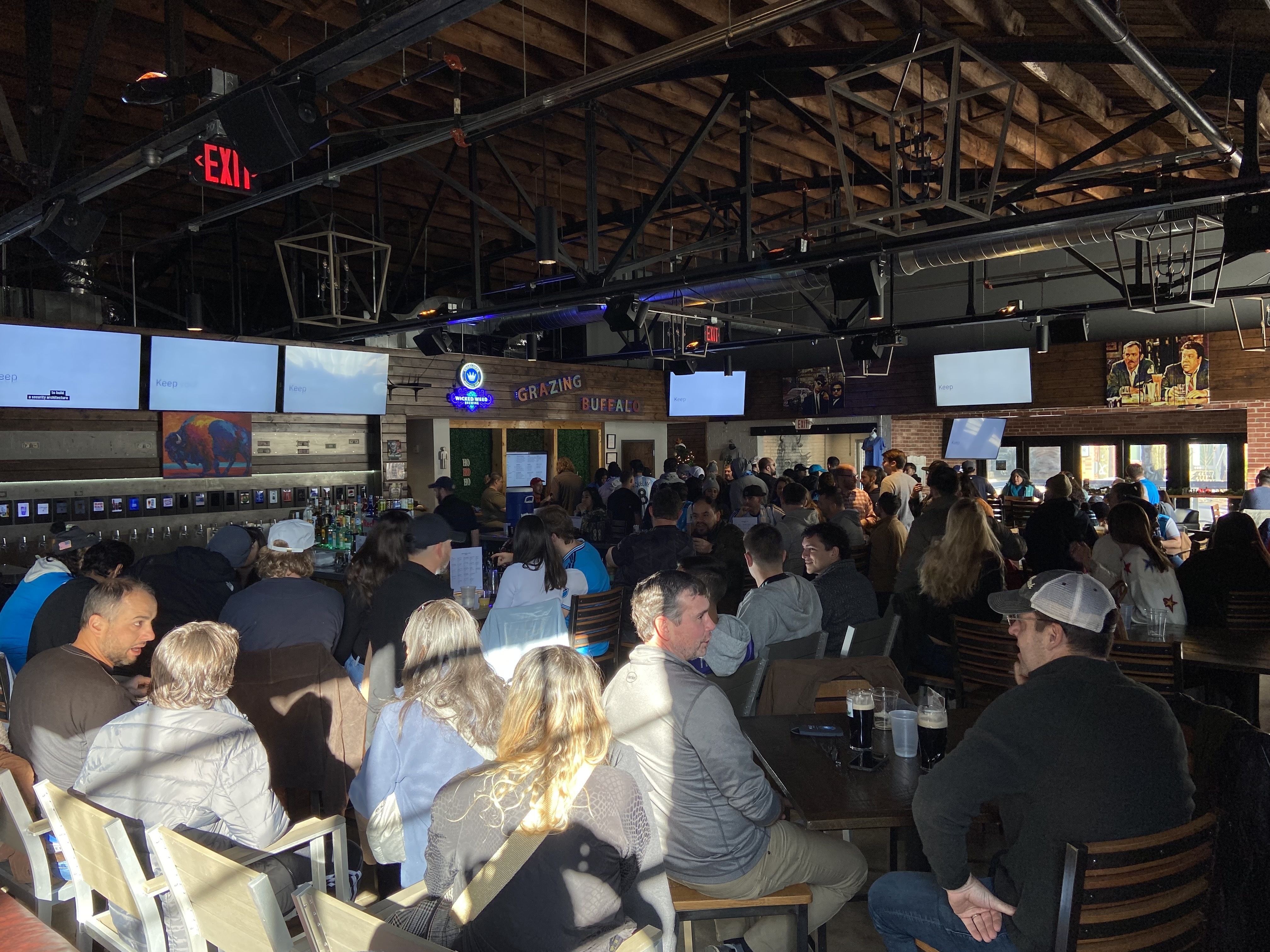 Busy indoor beer hall with exposed wooden beams and metal trusses. Long communal tables full of people, beer taps, wall screens, and a neon sign reading "GRAZING BUFFALO".