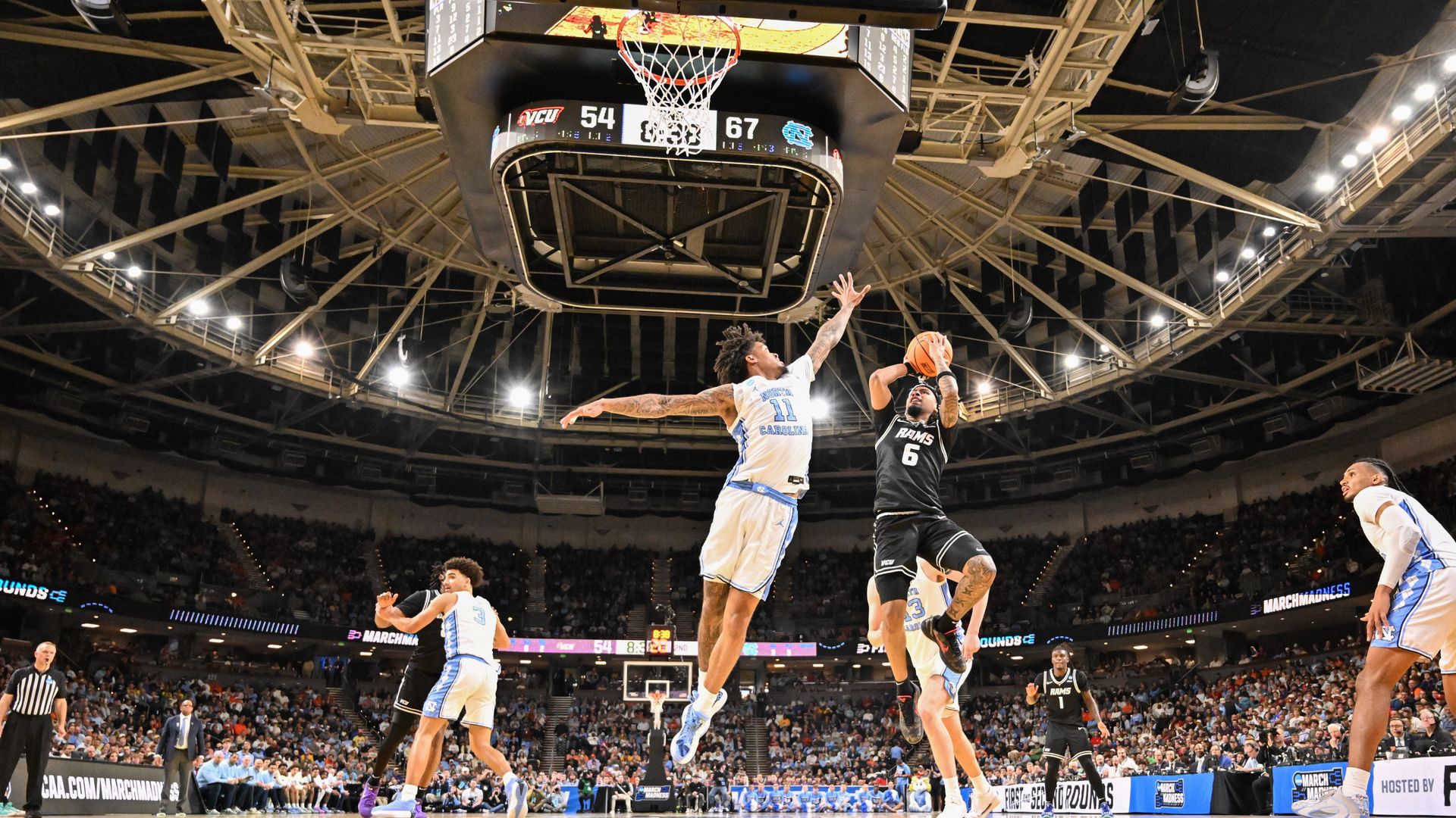 Basketball game in a packed arena: a white-and-blue team guard (No. 11) leaps to defend as a black uniformed player (No. 6) goes up for a shot near the basket; crowd and scoreboard visible.