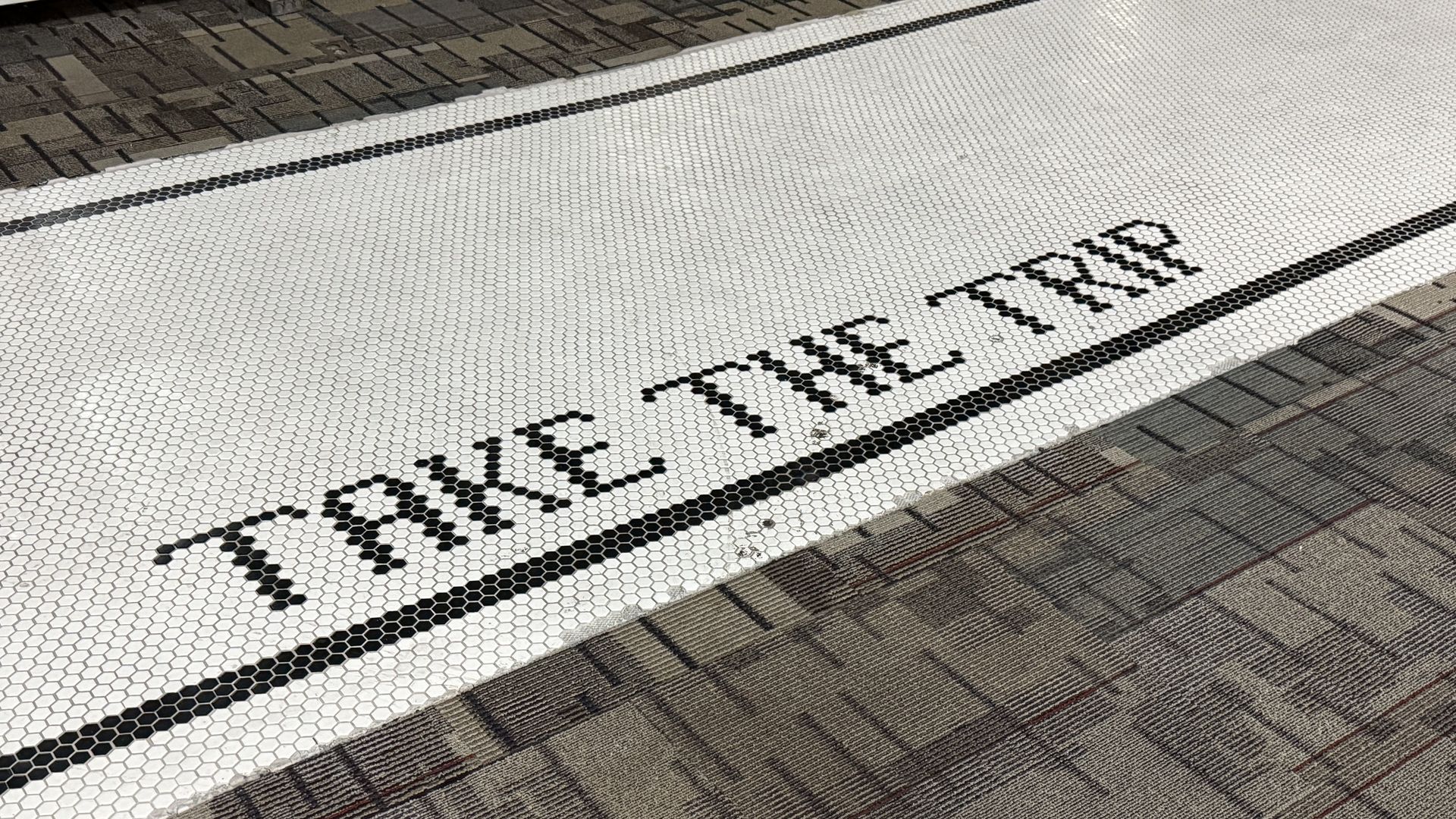 Close-up of a white hexagonal-tile strip on a sidewalk, bordered by a black line. Brown brick pavement frames both sides. Black letters spell a word along the edge of the tile strip, cropped.