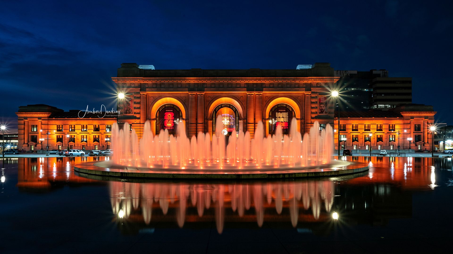 Night view of a historic building illuminated in orange lights with a large circular fountain in front, water jets glowing orange and reflecting on the still water surface.
