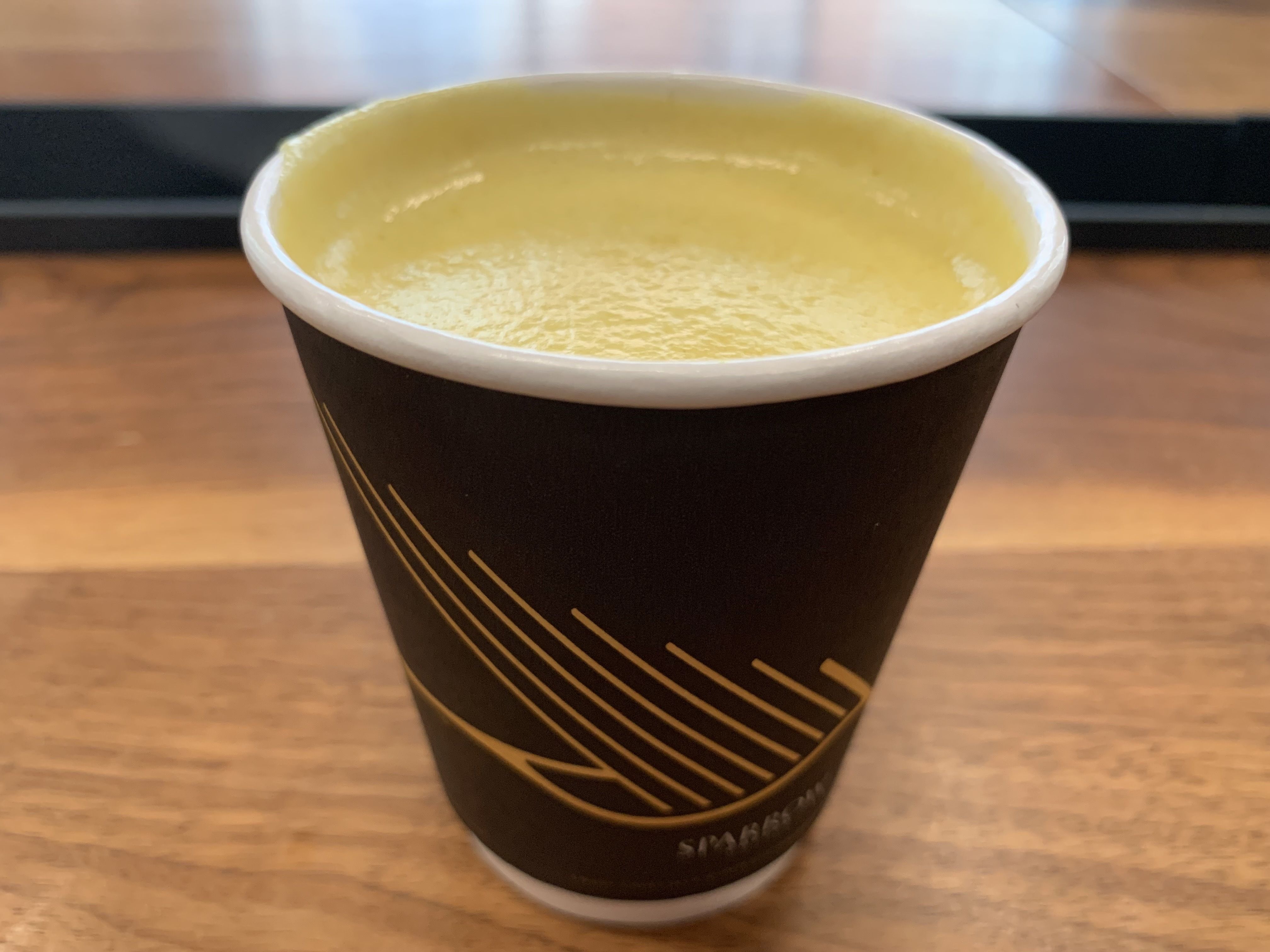 Top-down close-up of a black to-go coffee cup with a white rim and subtle sleeve, filled with pale yellow, foamy coffee, on a wooden table with a blurred dark background.