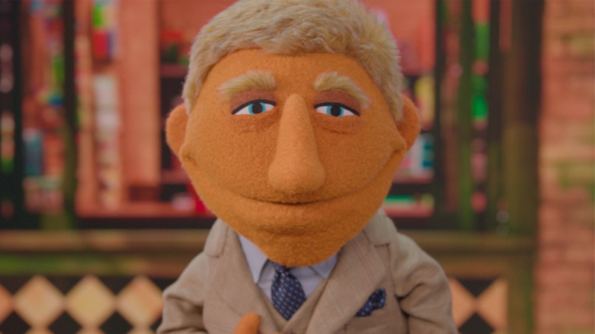 Puppet character with orange skin, blond hair, and blue eyes wearing a beige suit and blue tie, standing in front of a blurred colorful background.