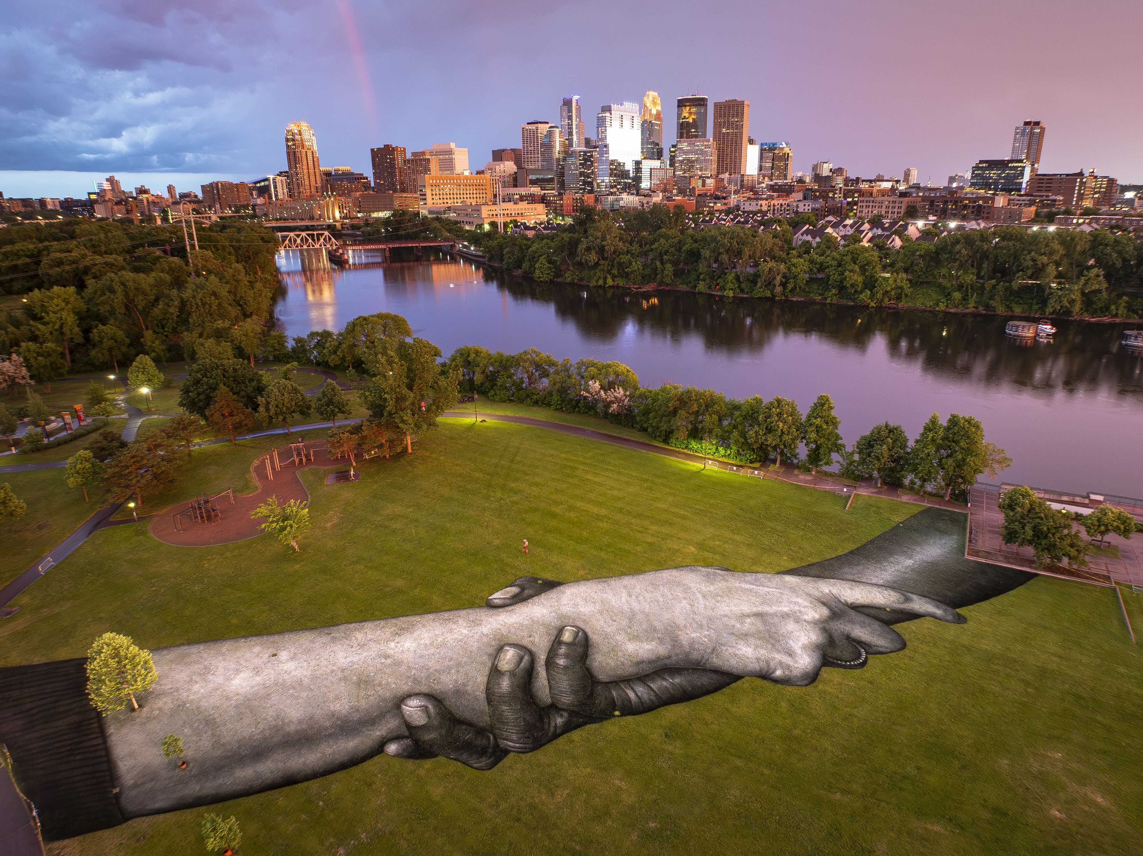 City skyline at sunset over a calm river, with a faint rainbow in a pink-purple sky. In the foreground a green park and a long dark sculptural installation lying across the grass with holding hands. 