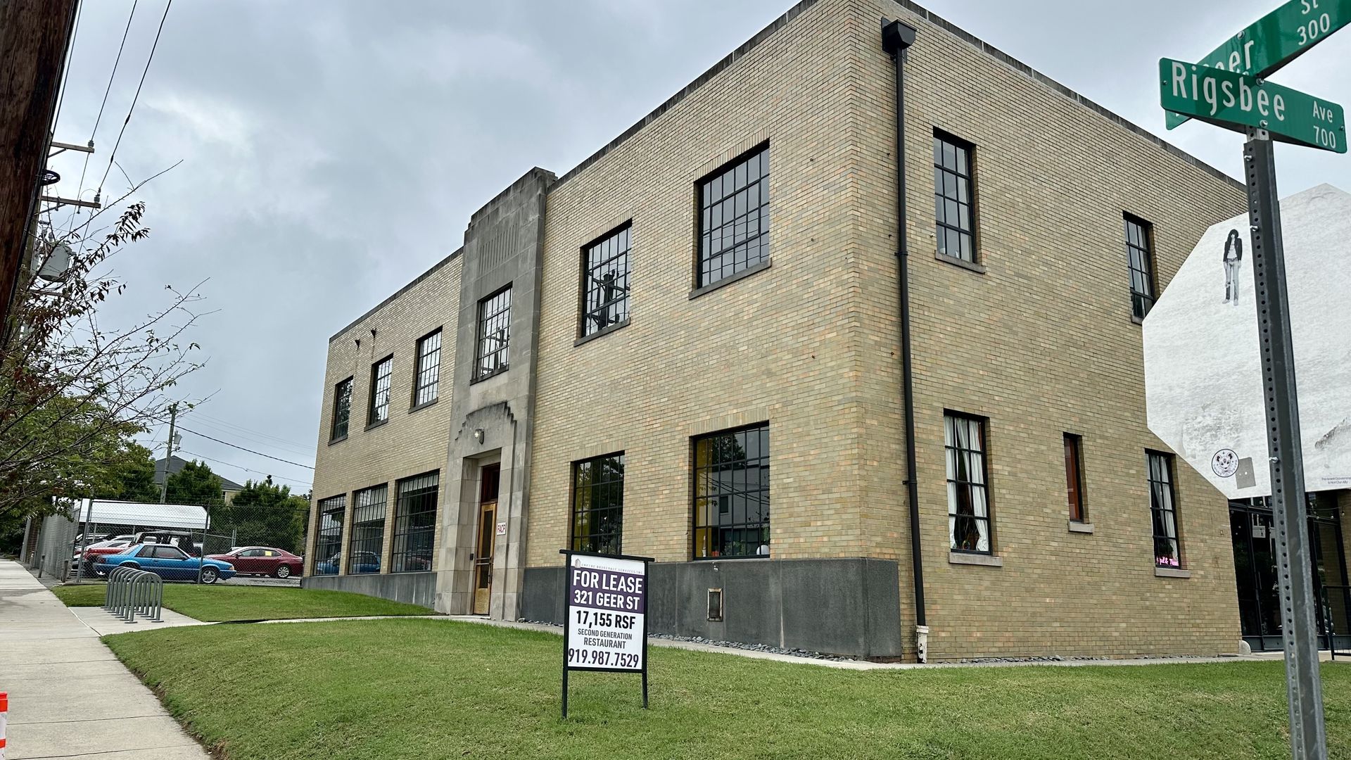 Overcast day at a street corner with a beige brick building, large black-framed windows, a green Rigsbee Ave street sign, a For Lease sign out front, and a utility pole on the left.