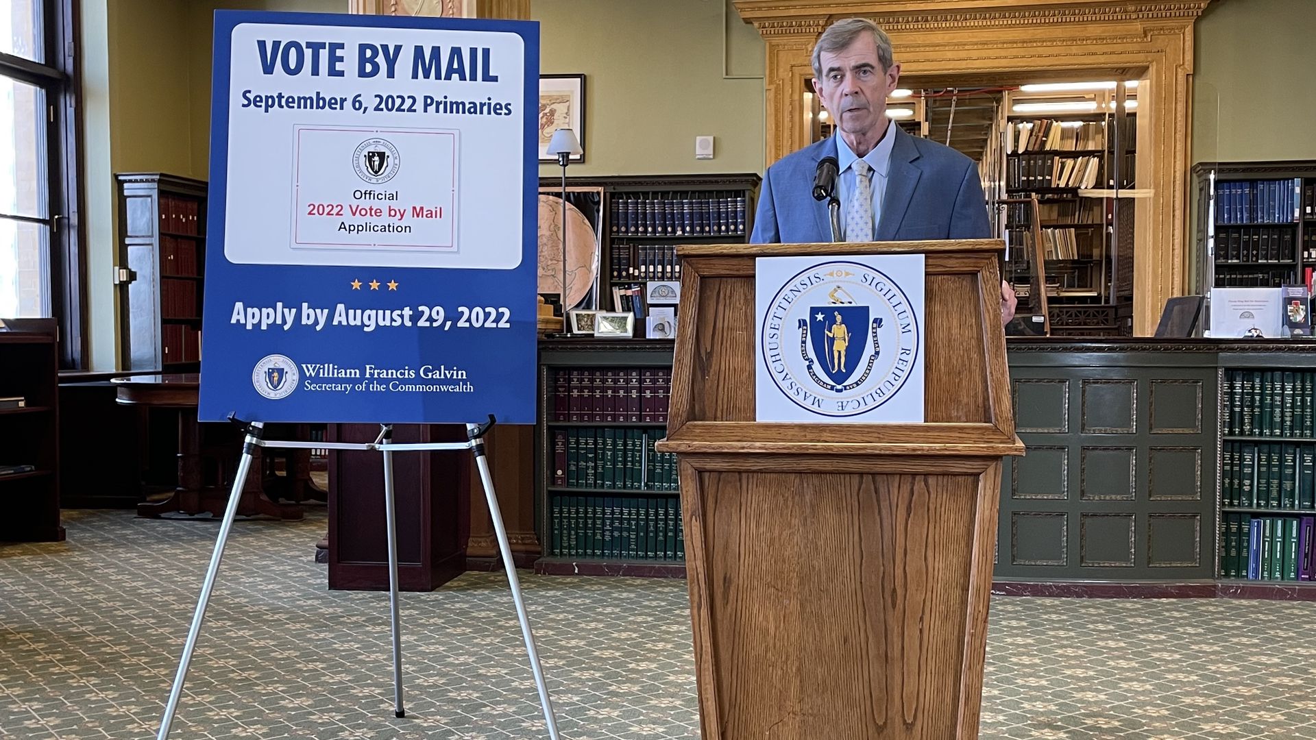 Secretary of State Bill Galvin stands behind a wooden podium next to a large blue and white "vote by mail" sign in the state library.
