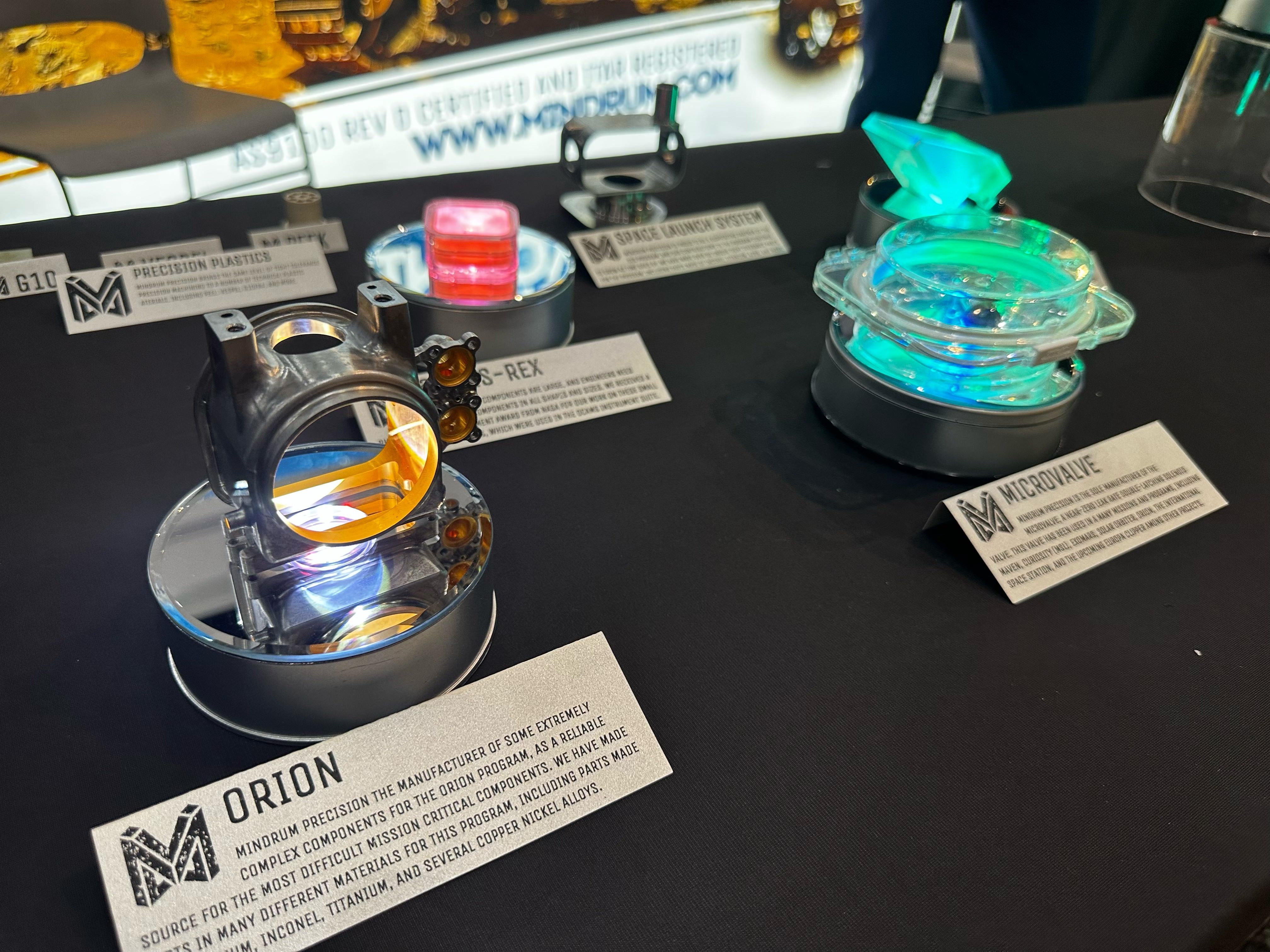 Exhibit table showing a chrome mechanical part with an orange interior beside a glowing turquoise circular device; labels read "Orion" and "Microvalve".