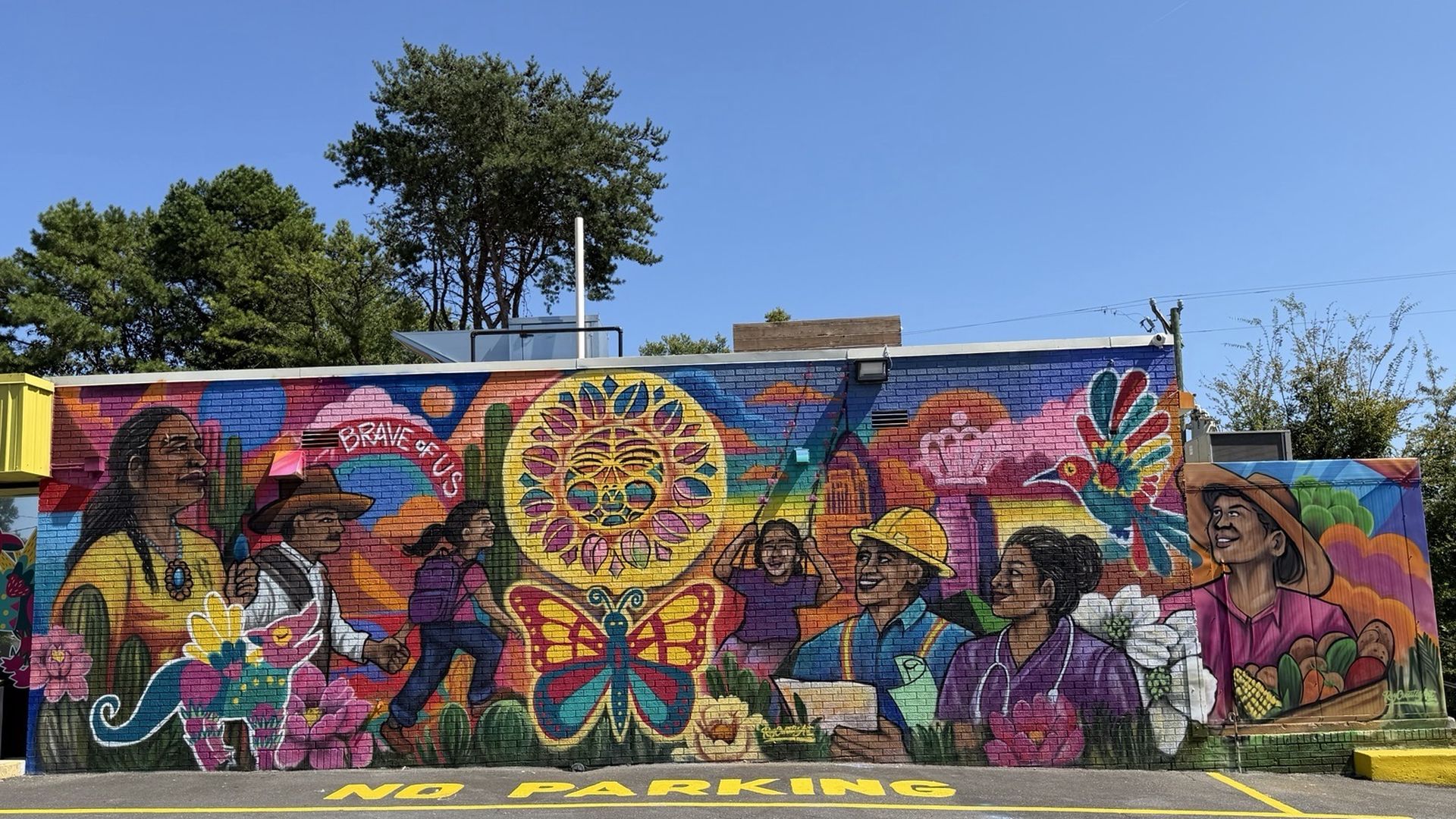 Vibrant mural on a brick wall shows diverse people dancing and smiling around a bright sun circle, with a butterfly, birds, and flowers; the pavement foreground reads "NO PARKING".