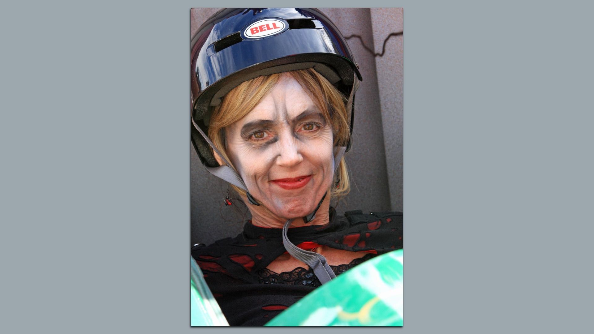 Close-up of a person wearing a glossy black Bell helmet with pale, zombie-like makeup, dark eye shadow, and red lipstick, against a gray textured wall.