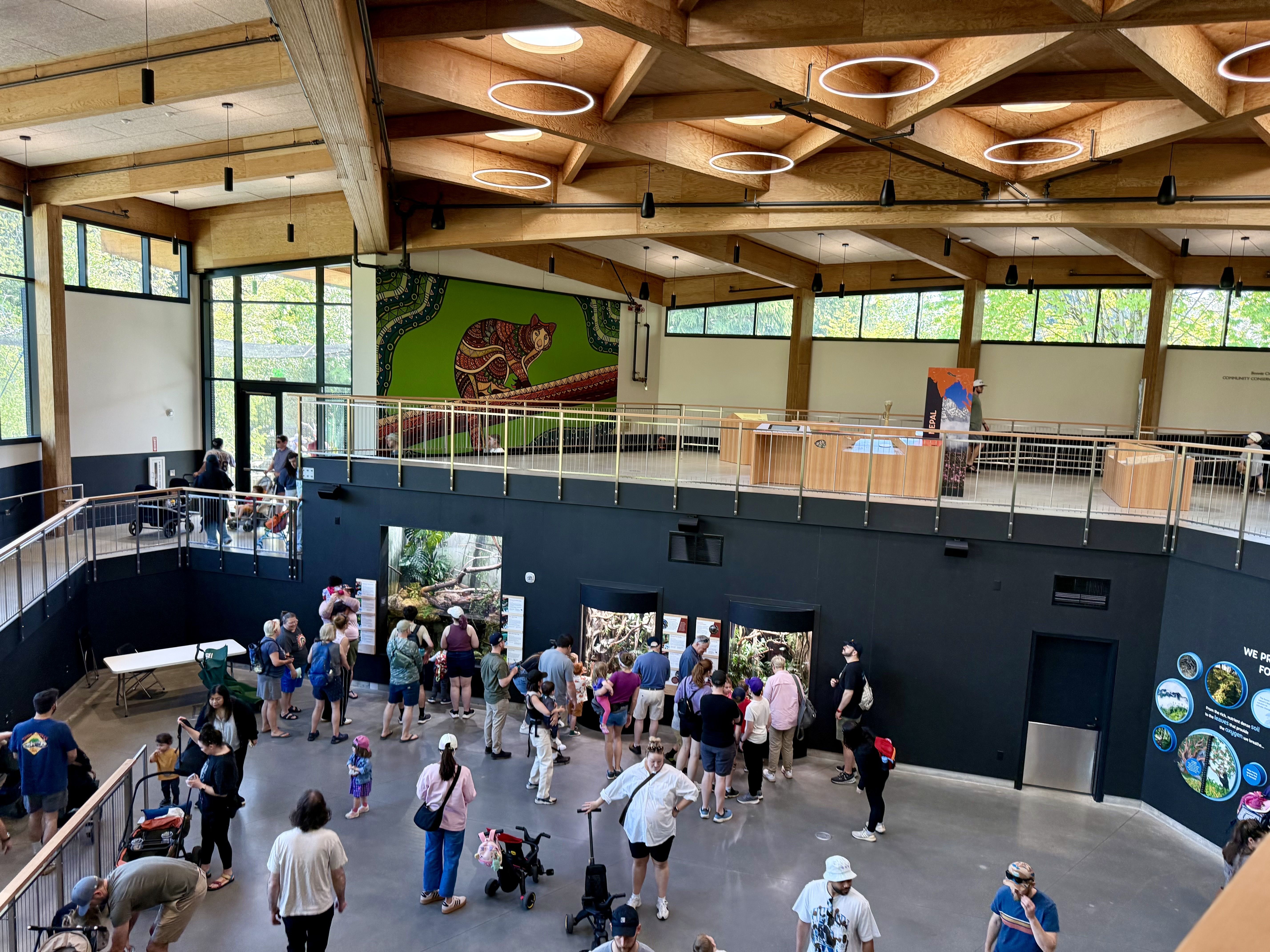 Indoor museum atrium with exposed wooden beams and circular ring lights. A mezzanine balcony overlooks a crowded main floor of visitors; a large green mural features a stylized patterned animal.