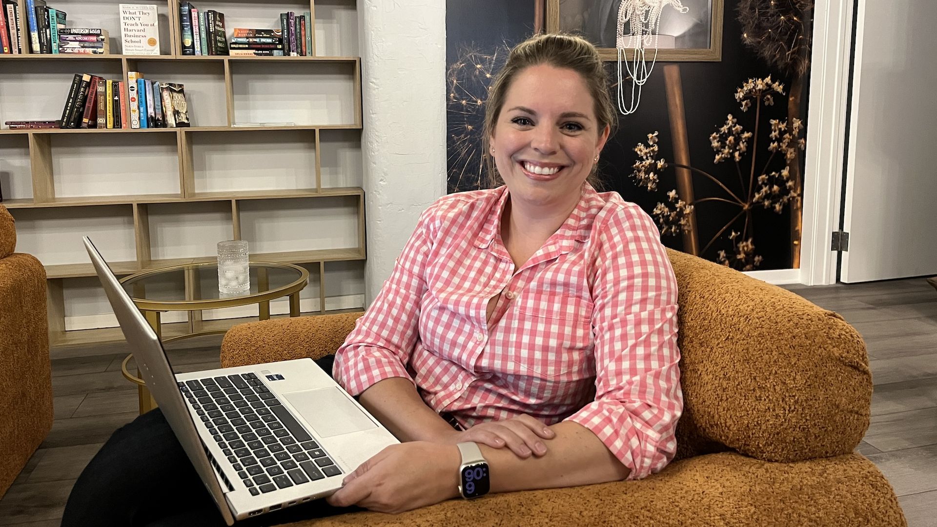 Woman in pink checked shirt sitting in an orange chair with a laptop on her lap.