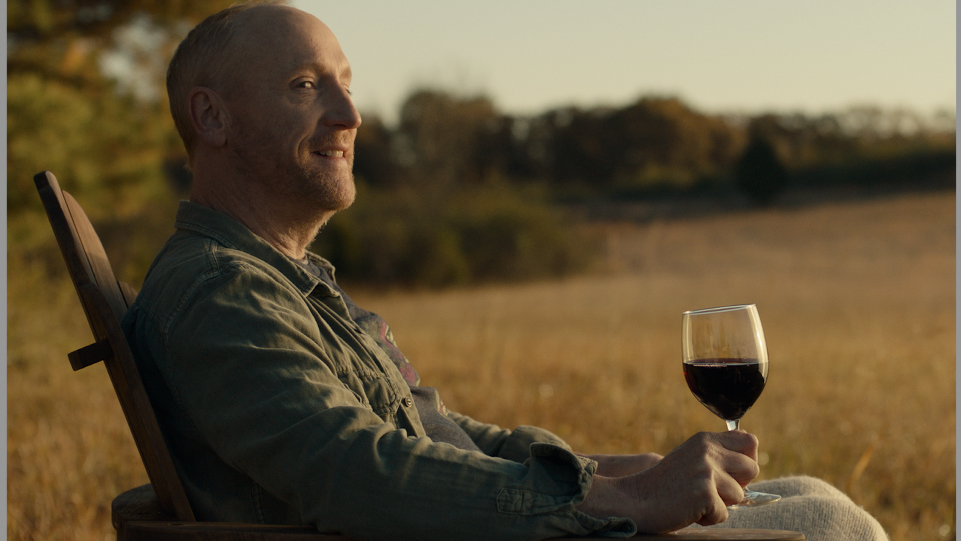 Production still featuring an actor sitting in a chair drinking a glass of wine.