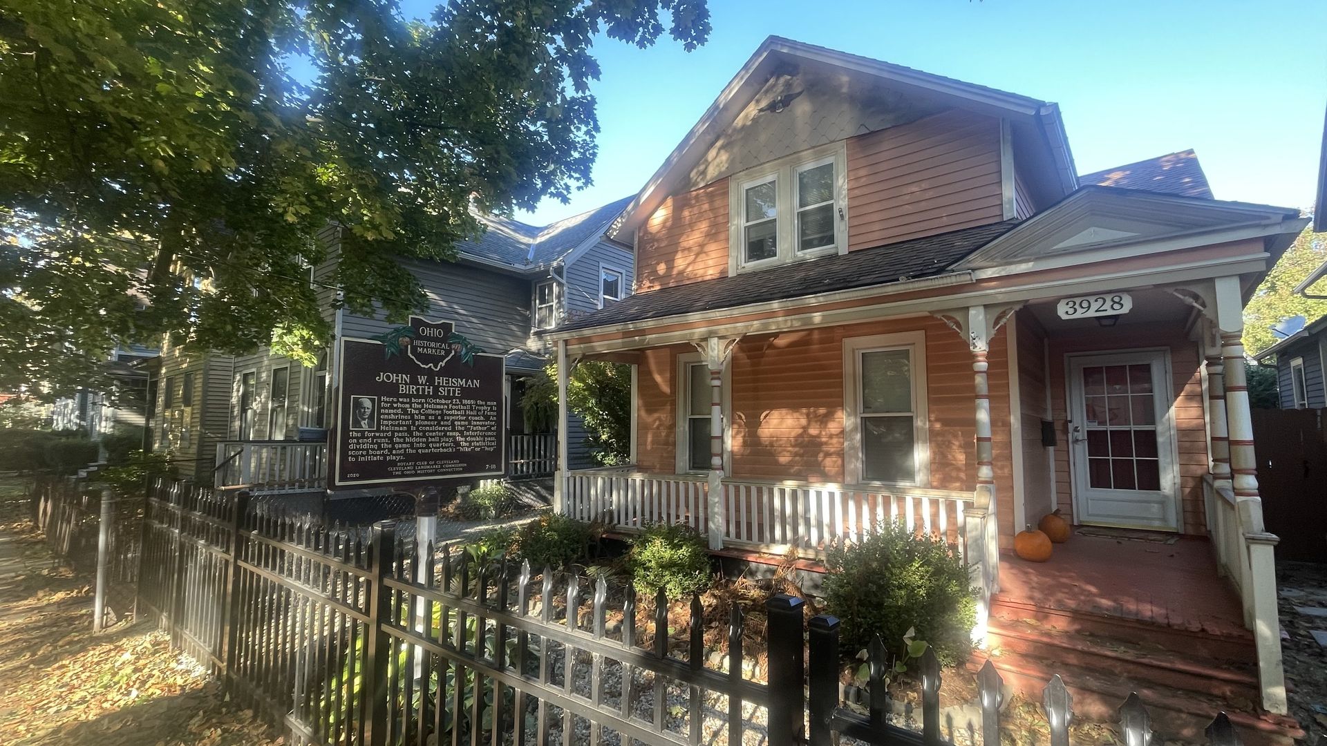 Sunlit street view of 3928, a brown-orange two-story house with white trim and a porch. A marker reads "John W. Heisman Birth Site" beside a white picket fence, under a leafy tree; pumpkins near the door.