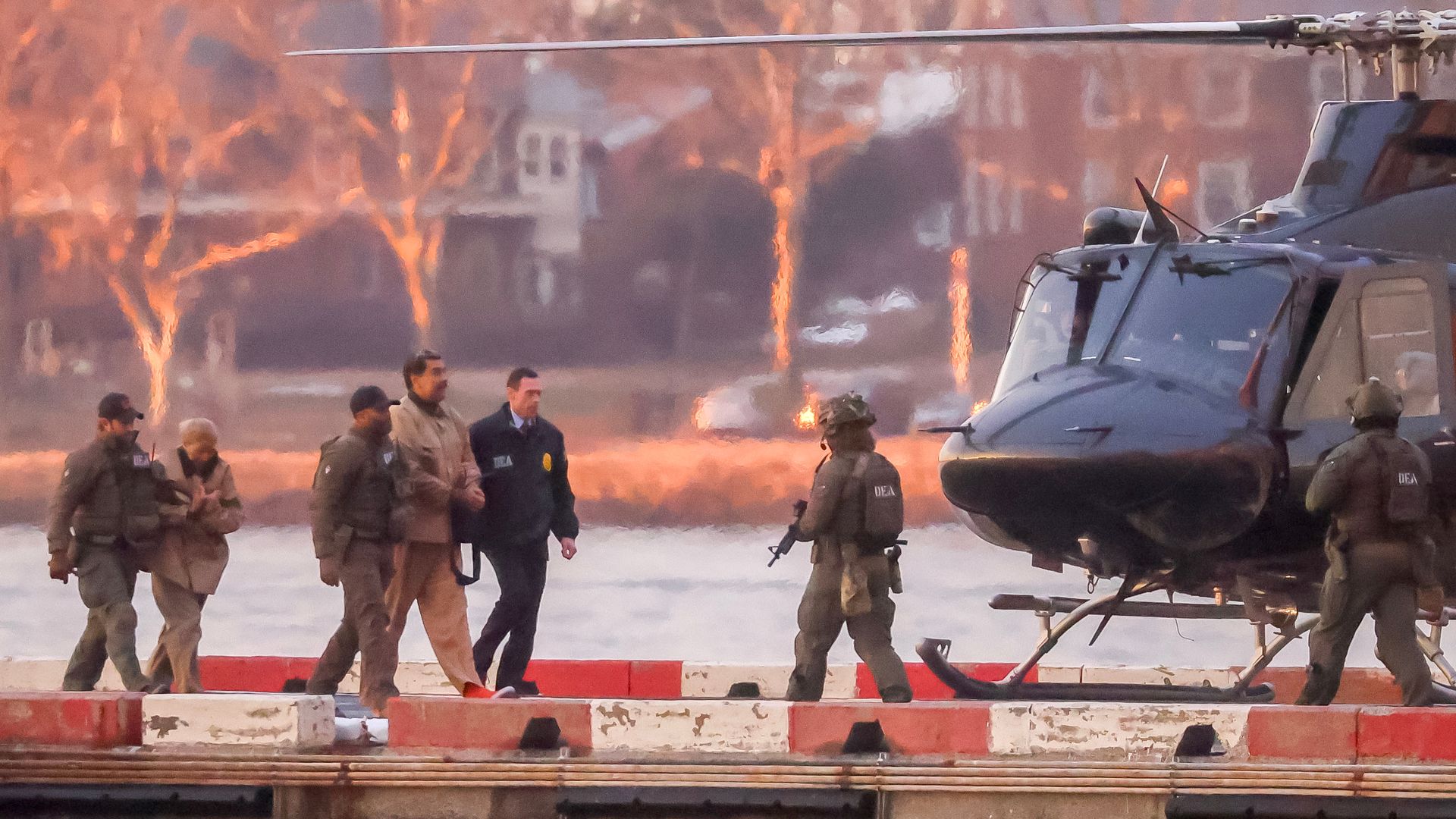 A military helicopter on a runway with soldiers walking in front; orange flames and smoke rise in the blurred urban background, suggesting a battlefield.
