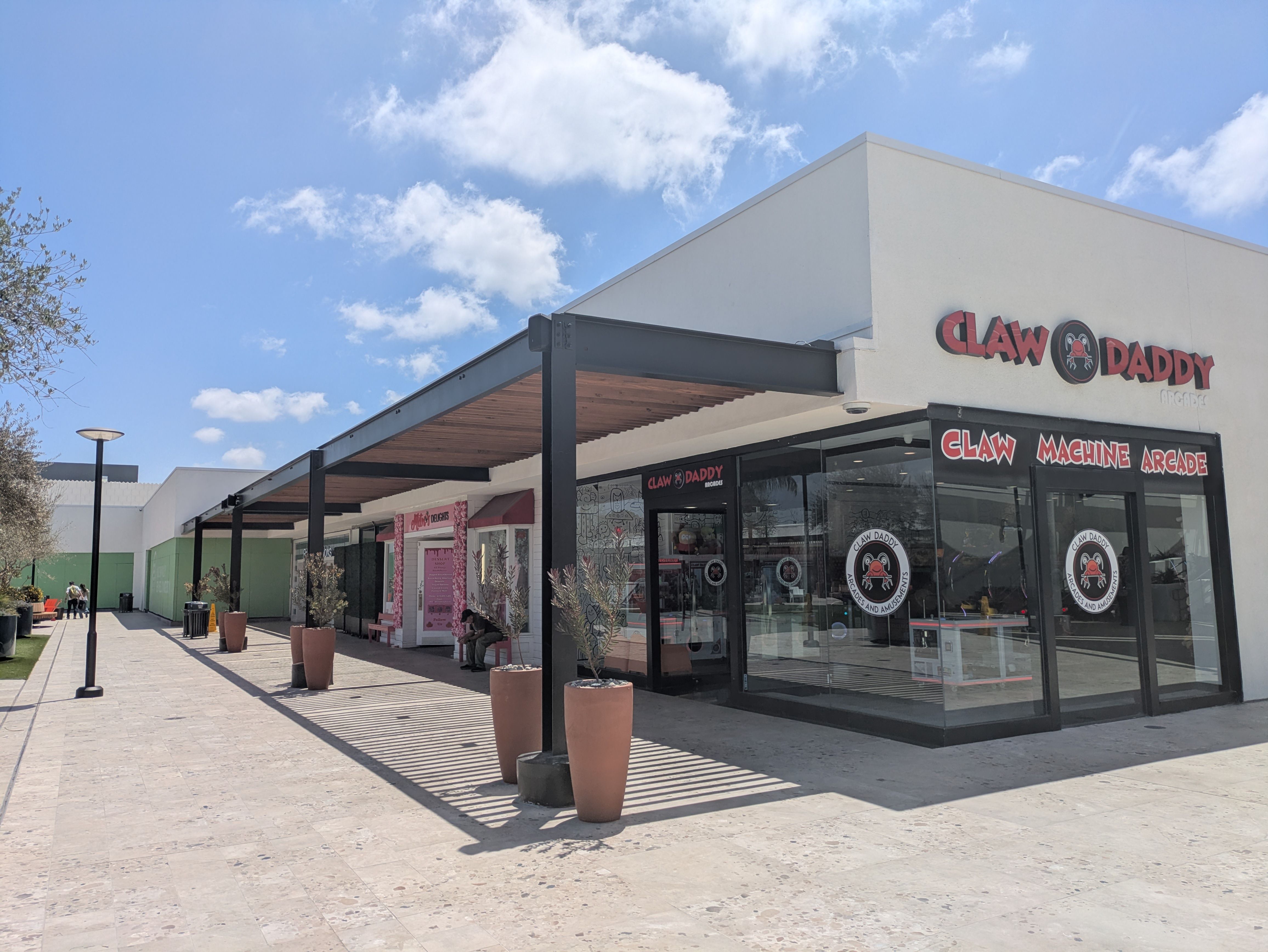 Bright day at a shopping complex with a white building, glass storefronts, and a red sign reading "Claw Daddy" with a crab logo; a covered walkway, potted trees, and a blue sky with clouds.