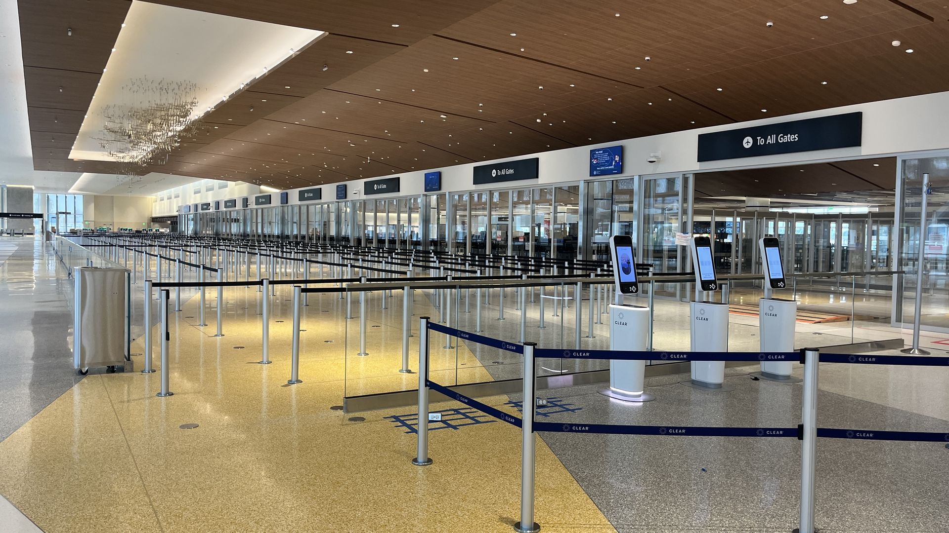 Empty airport security checkpoint area with numerous stanchions and black belts guiding lines, white CLEAR kiosks, wood-paneled ceiling, and signs saying "To All Gates."