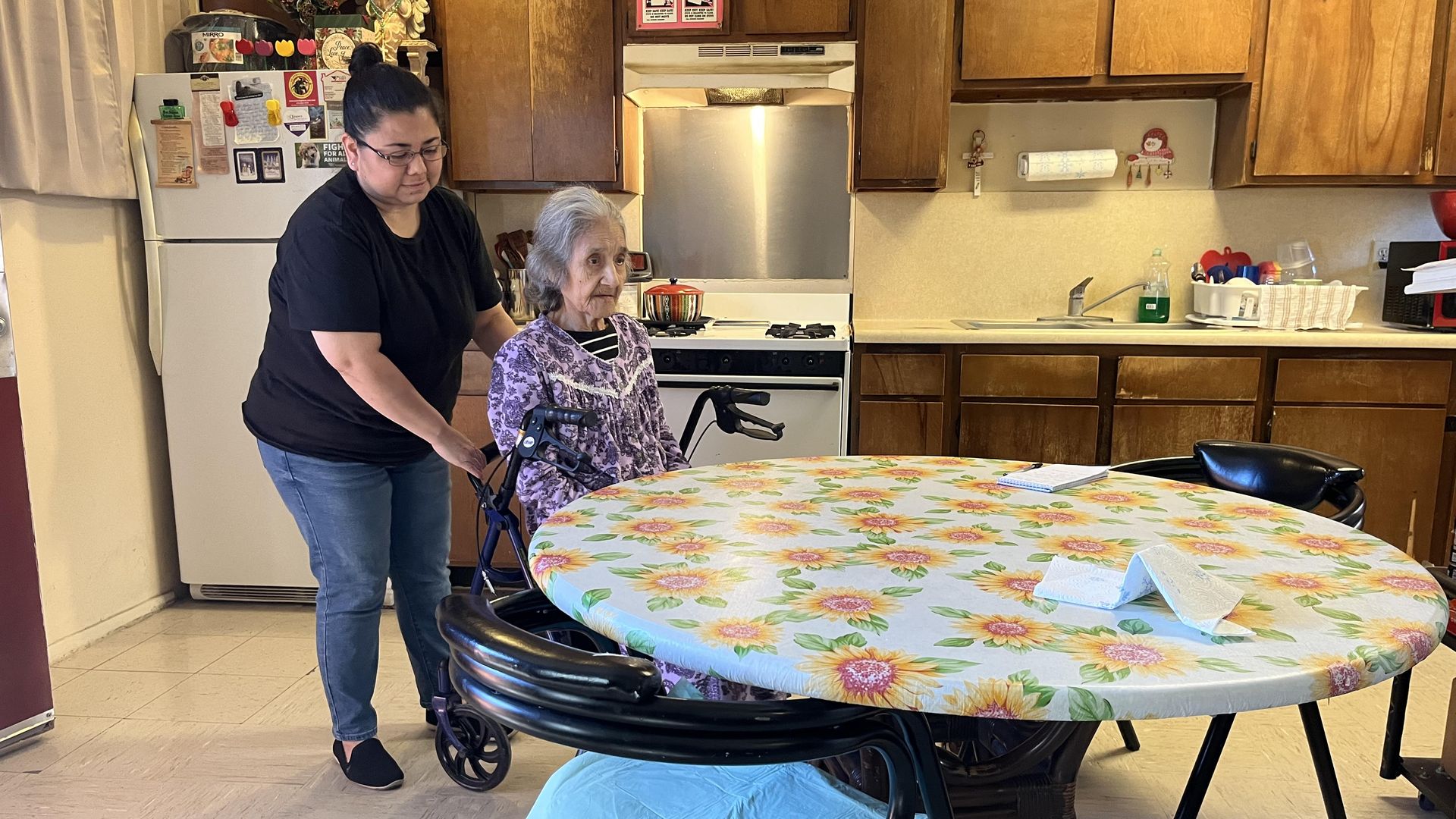 A woman helps an older women in a wheelchair sit down at a kitchen table.