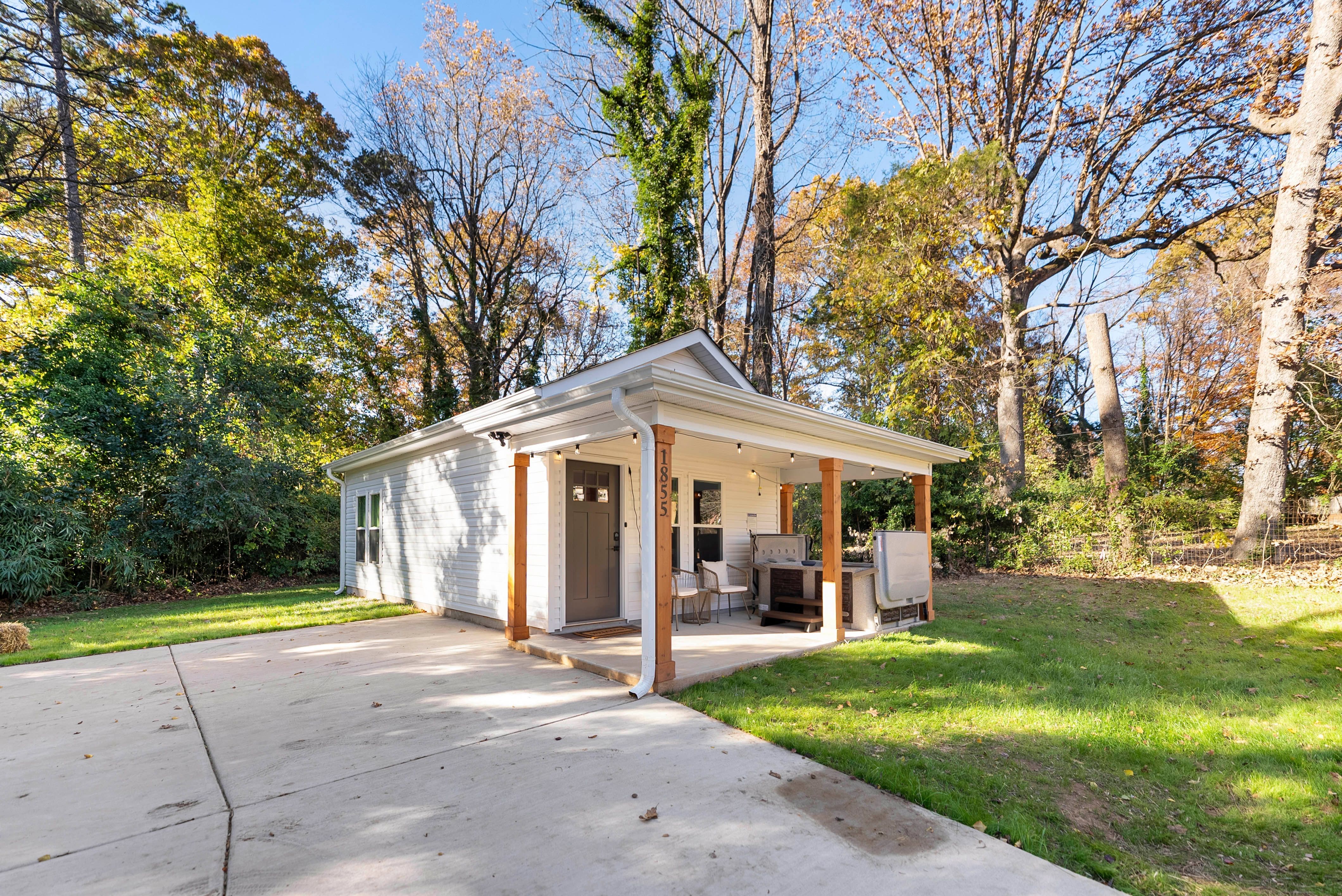 Small white house with a covered porch supported by wooden posts, gray door, two chairs, surrounded by green grass and tall trees with autumn foliage under a clear blue sky.
