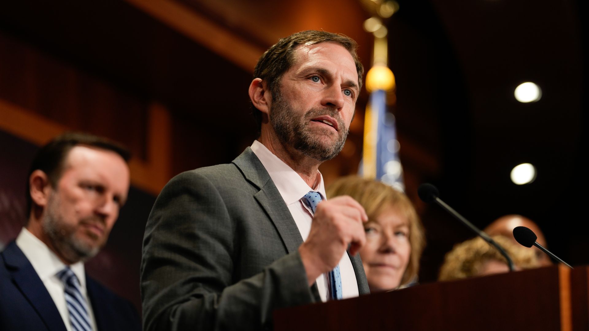 Rep. Jason Crow, wearing a gray suit and speaking at a wooden podium flanked by colleagues in a wooden room.