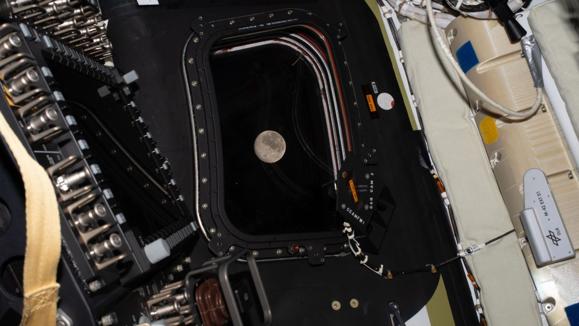 Interior of a spacecraft with a black rectangular hatch frame; the Moon floats near the opening, surrounded by cables, panels, and beige insulation.