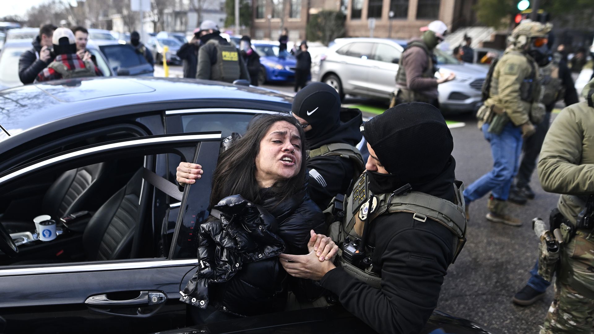 Two immigration enforcement agents wearing vests and black jackets with hoods pull up a woman from her car.