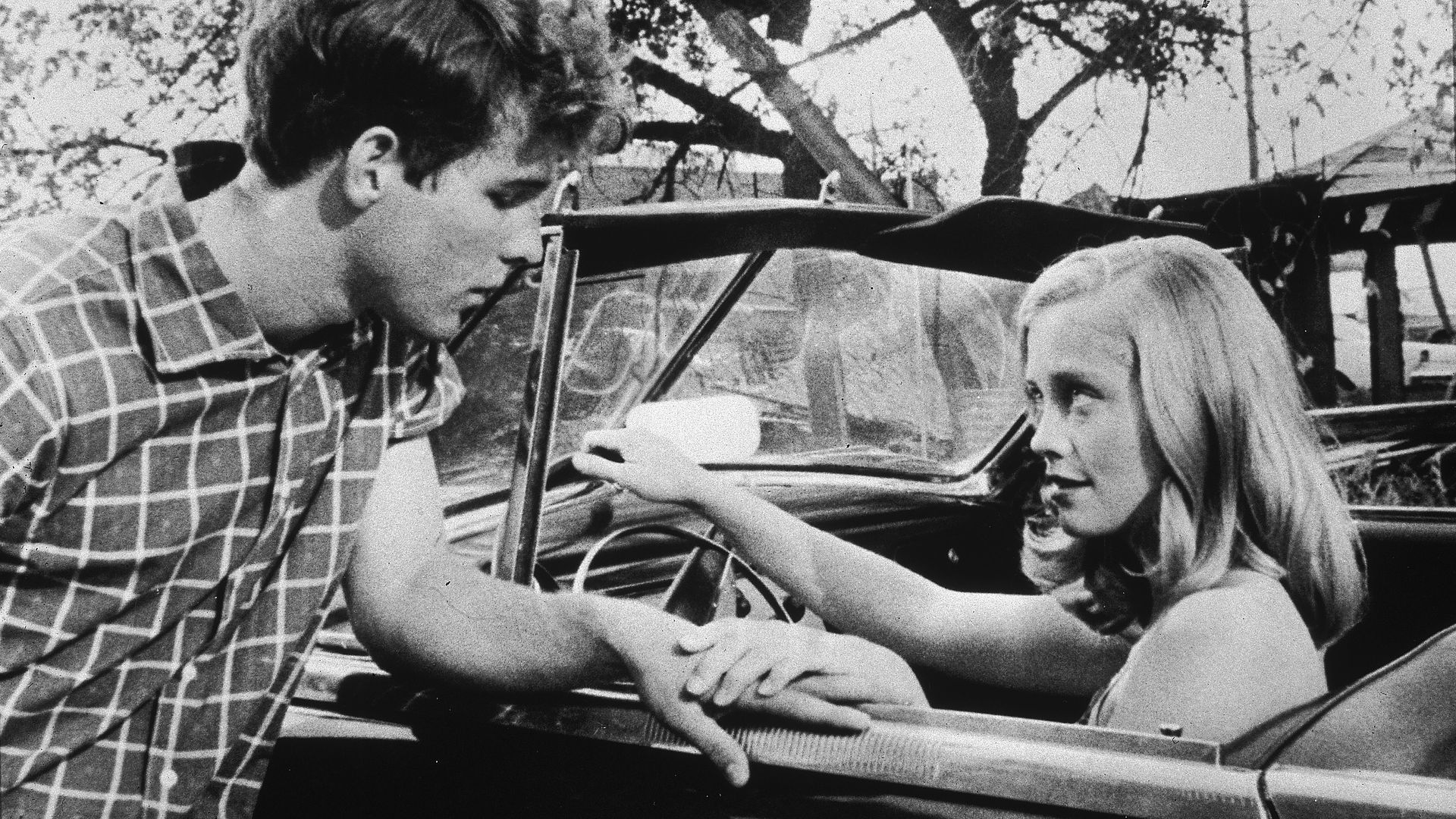Black-and-white photo of a young man in a checkered shirt and a girl with long hair beside a vintage convertible outdoors under trees; she touches the car door as he looks down.