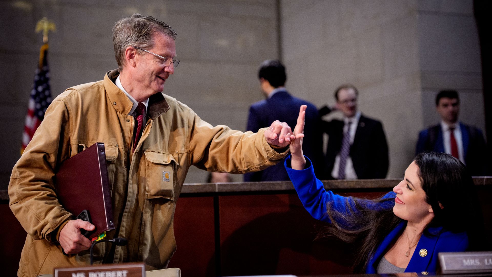 A man in a tan jacket and red tie smiles as he touches fingers with a woman in a blue blazer seated at a desk, with an American flag and other people blurred in the background.