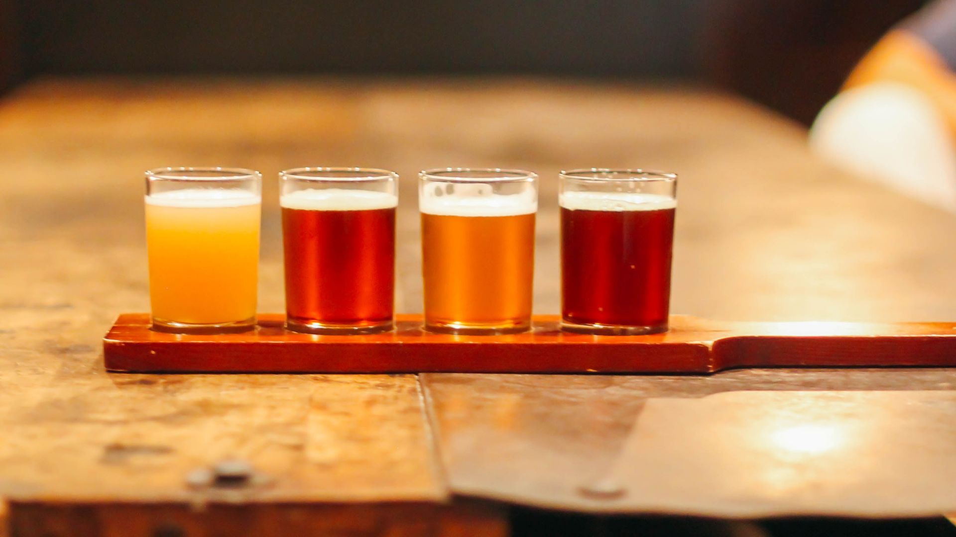 Four small beer tasters on a wooden flight paddle sit on a bar counter; left to right: pale ale, amber, golden, and dark red beers, with a blurred background.