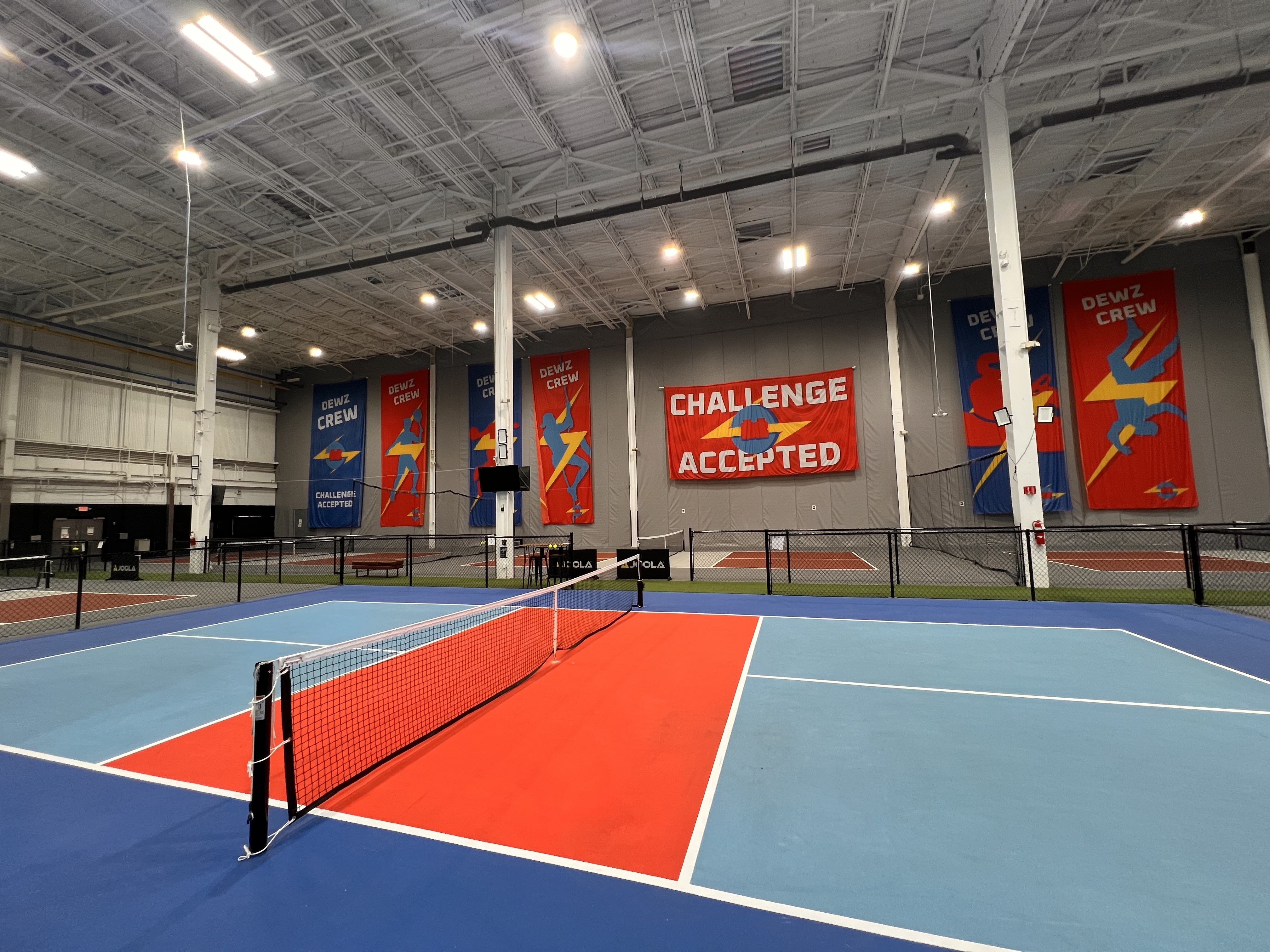 Indoor tennis facility with blue and red courts, nets, and a large red banner reading "CHALLENGE ACCEPTED." Blue and red banners with stylized athletes line the walls, while bright overhead lights illuminate the space.