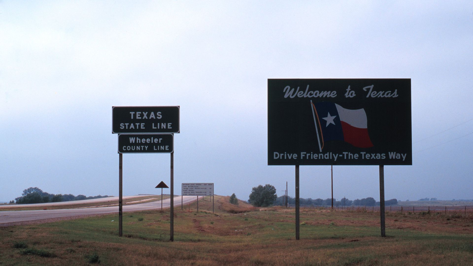 Road signs in a rural area: one reads "Texas State Line Wheeler County Line", another says "Welcome to Texas" with a waving Texas flag and the phrase "Drive Friendly-The Texas Way."