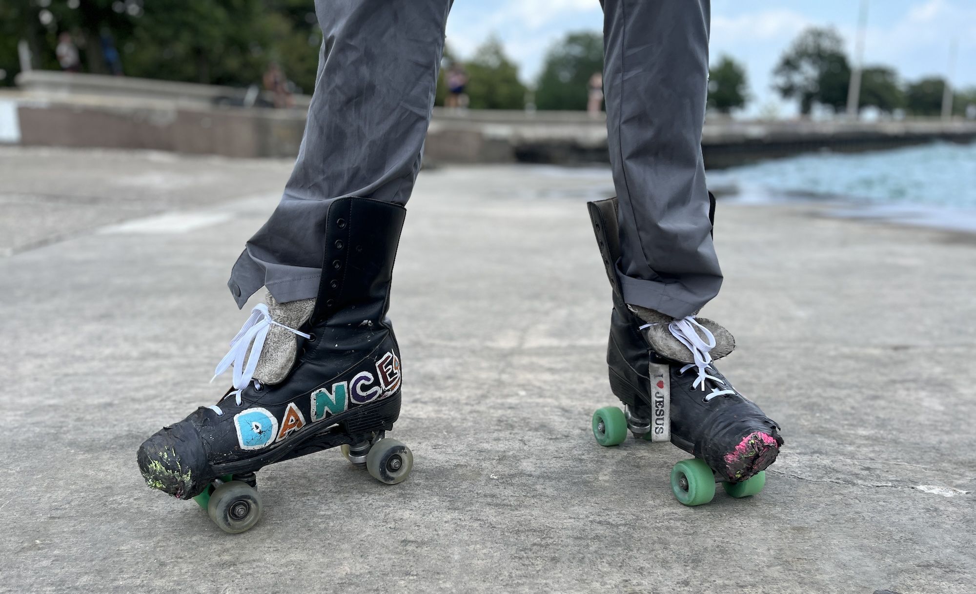 Legs wearing gray pants and black roller skates with white laces, standing on concrete near water. One skate has colorful letters spelling "DANCE"; the other has a tag reading "I ❤️ JESUS."