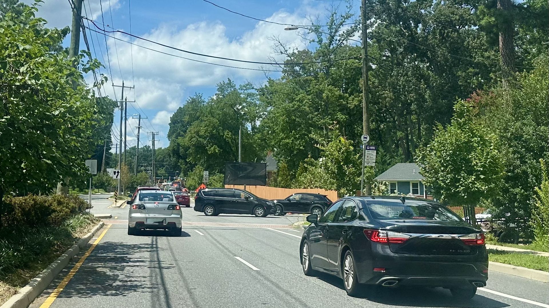 Sunny suburban street with parked and moving cars, tall trees on both sides, utility poles and power lines, a house visible on the right, blue sky with clouds.