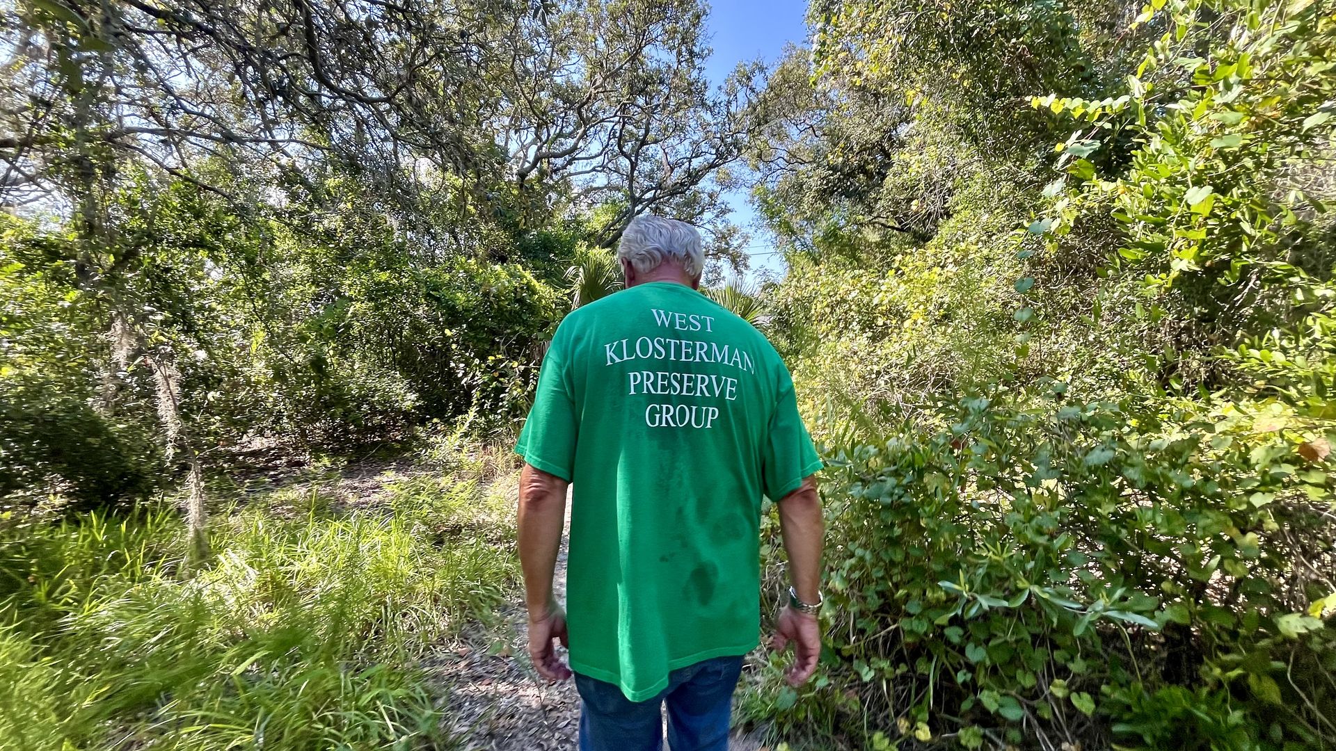 A man with white hair in a green shirt that reads "West Klosterman Preservation Group" on the back walks on a nature trail surrounded by lush greenery.