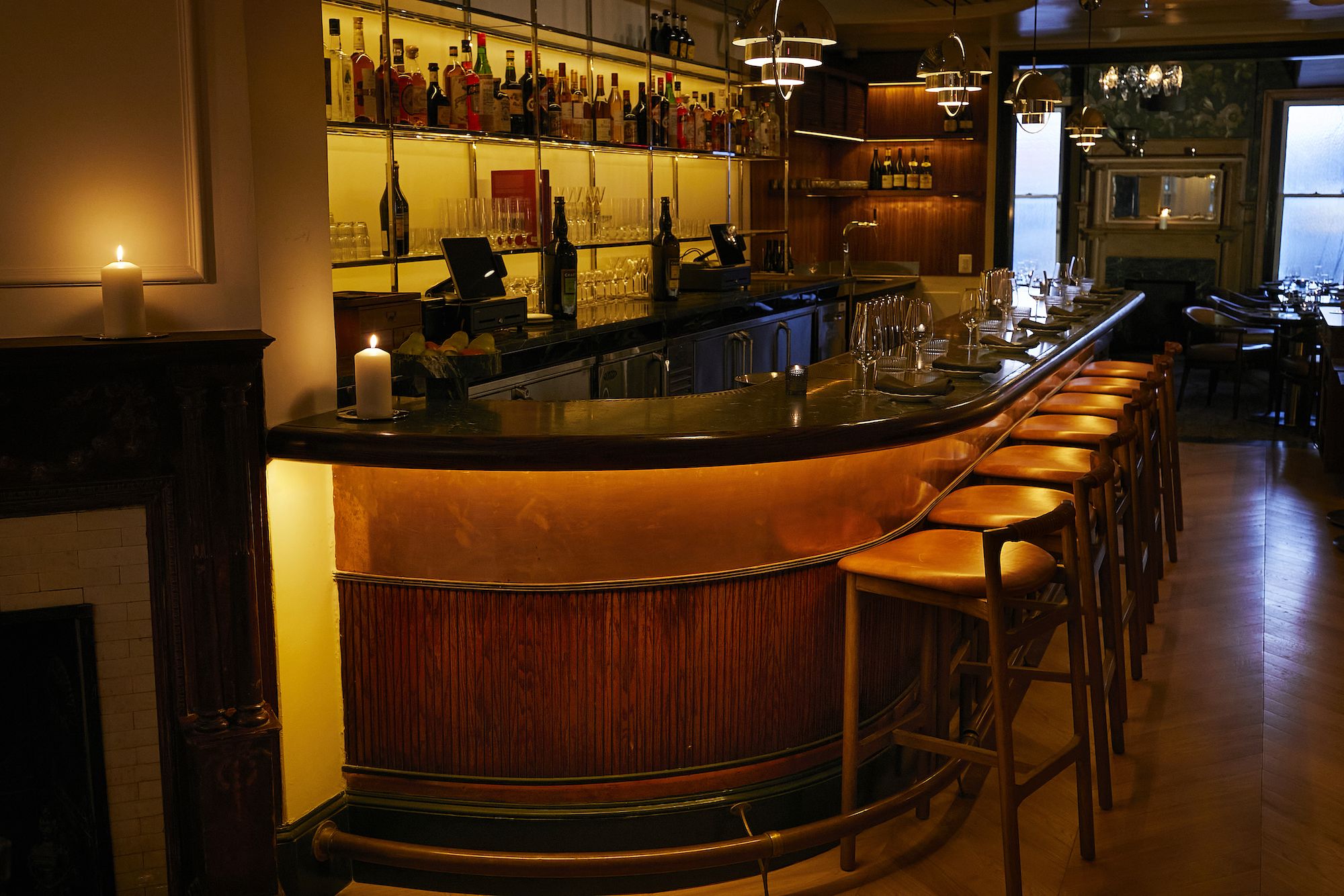 Warmly lit bar with amber backlighting and wooden stools with orange seats. Two candles glow on the bar counter, which is set with glasses and napkins. Shelves with bottles and glassware line the wall.